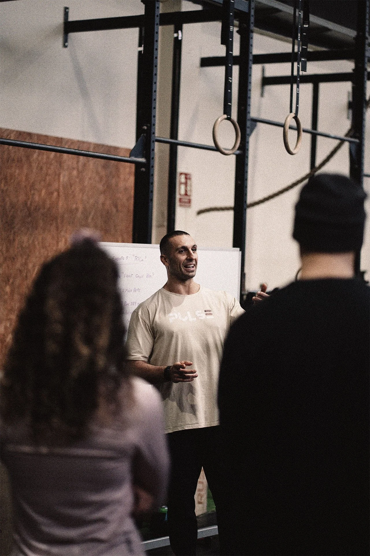 Un instructor da una clase en un gimnasio, frente a un grupo de personas que lo escuchan. Hay anillos de gimnasia y cuerdas en la estructura del gimnasio al fondo.