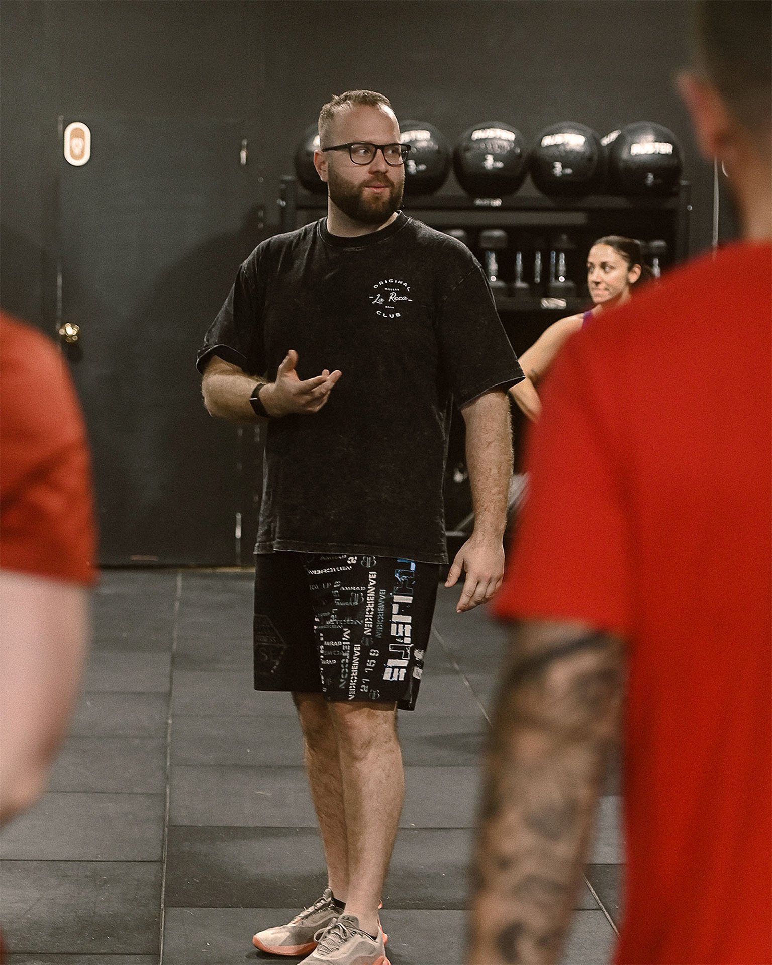 Un entrenador de gimnasio hablando con varias personas en un entrenamiento, en un gimnasio con pesas y balones medicinales en el fondo.