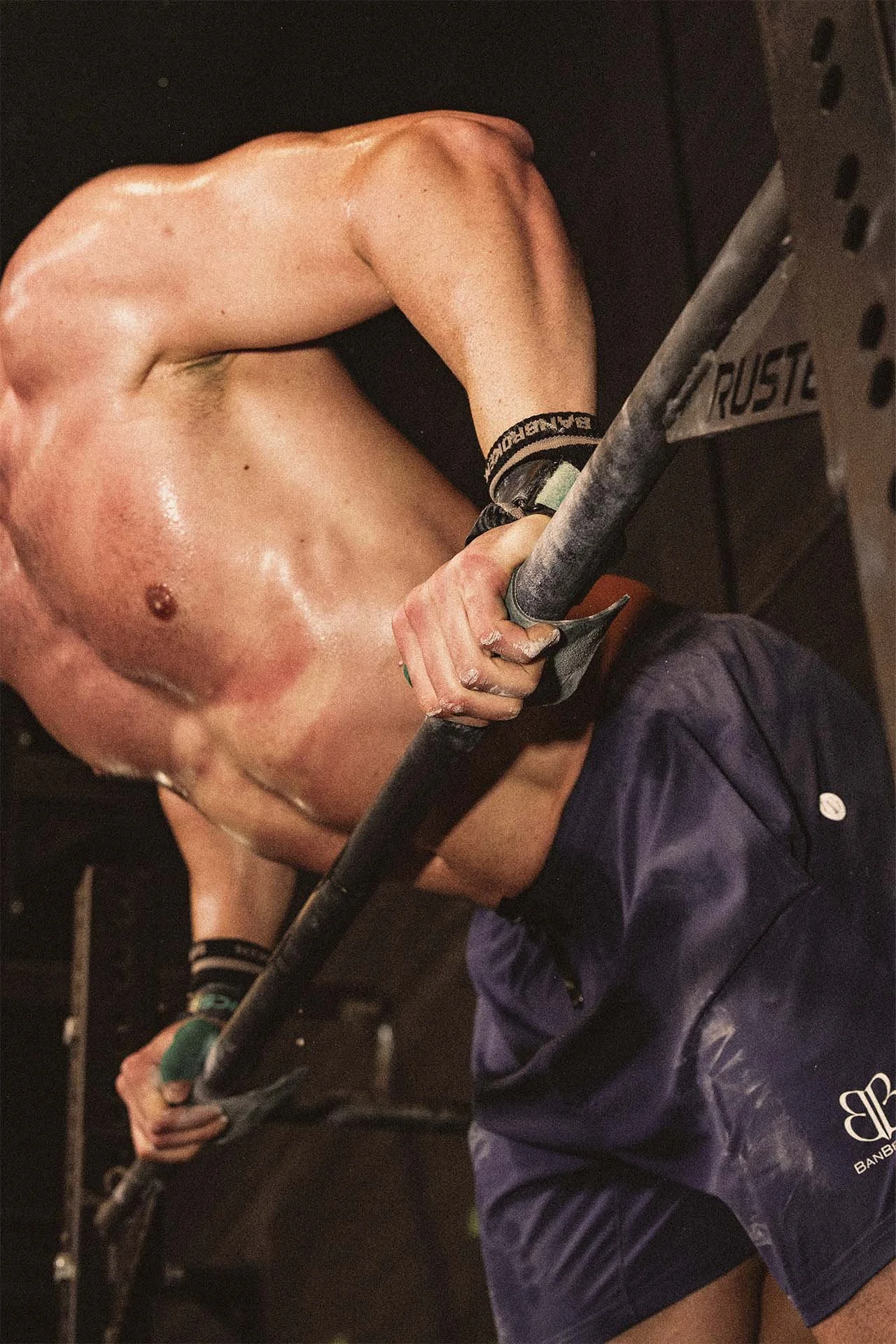 Persona realizando ejercicio de press de banca en un gimnasio, con pantalones cortos y sin camiseta, sudorosa, sujetando la barra con ambas manos.