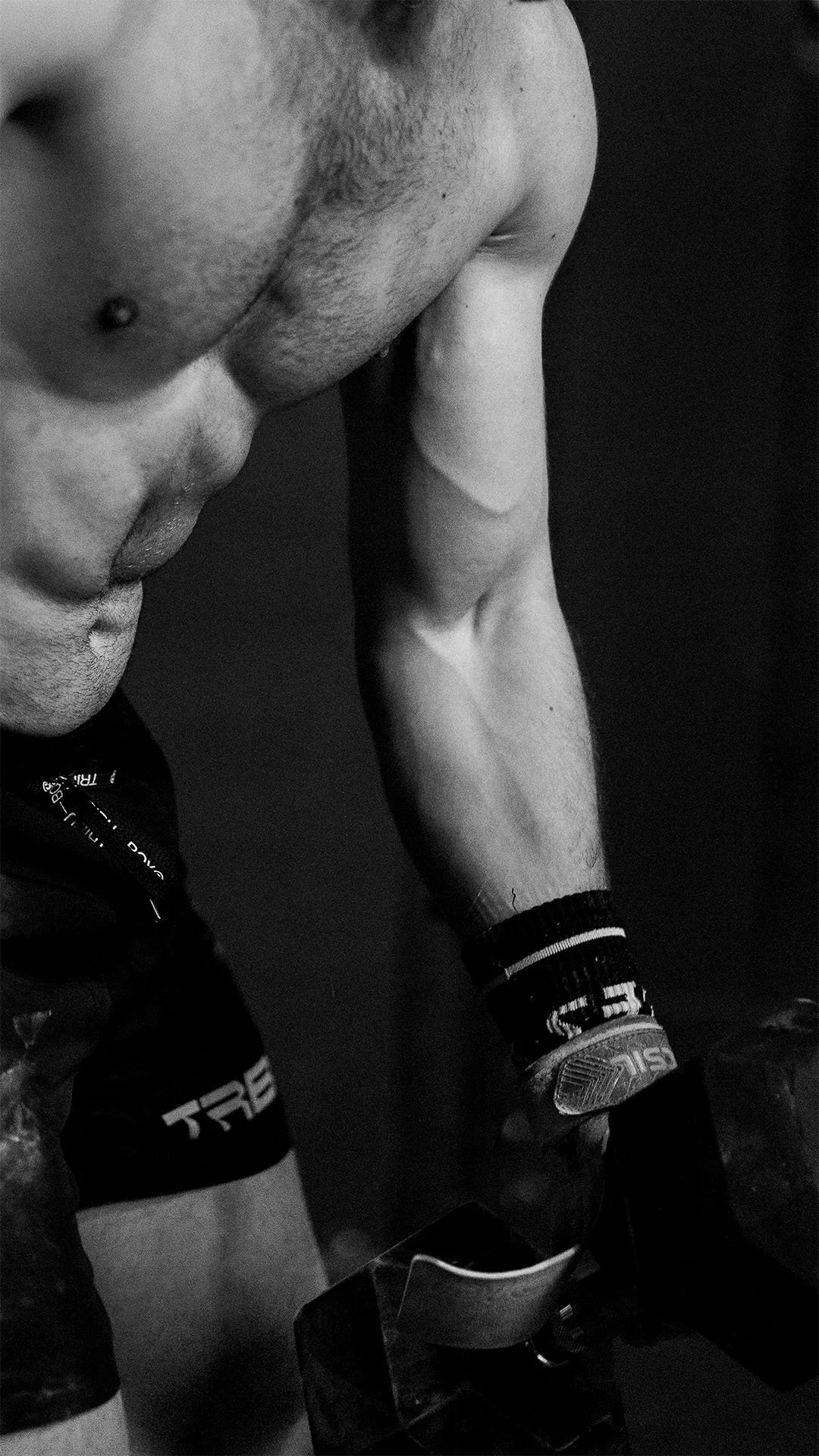 Una persona con una camisa deportiva, que lleva una pulsera y un reloj, está posando con el brazo doblado, mostrando su músculo. La imagen es en blanco y negro.