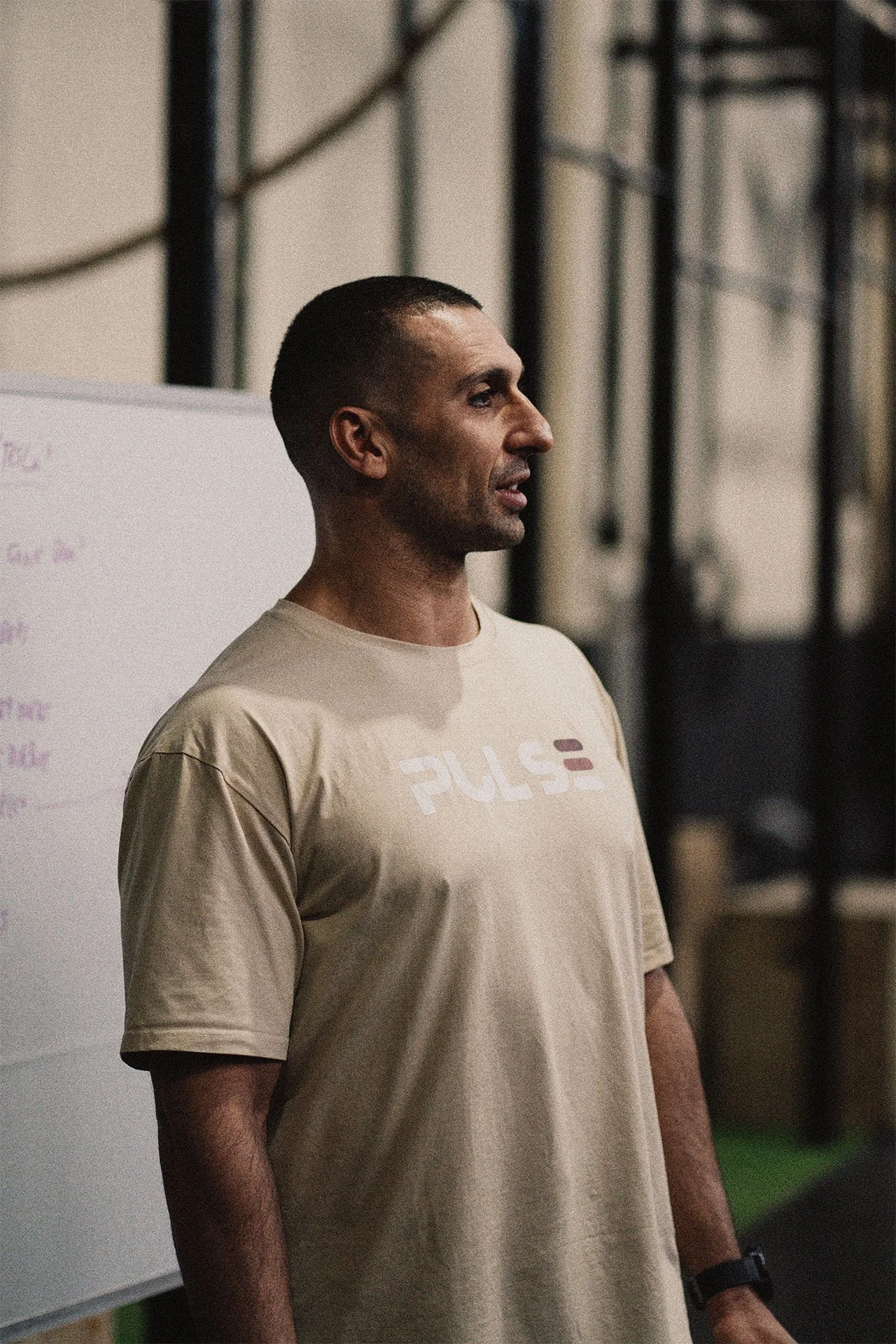 Hombre en un gimnasio con camiseta beige con la palabra 'PULSE' en la parte frontal, que parece estar dando una instrucción o hablando frente a una pizarra blanca.