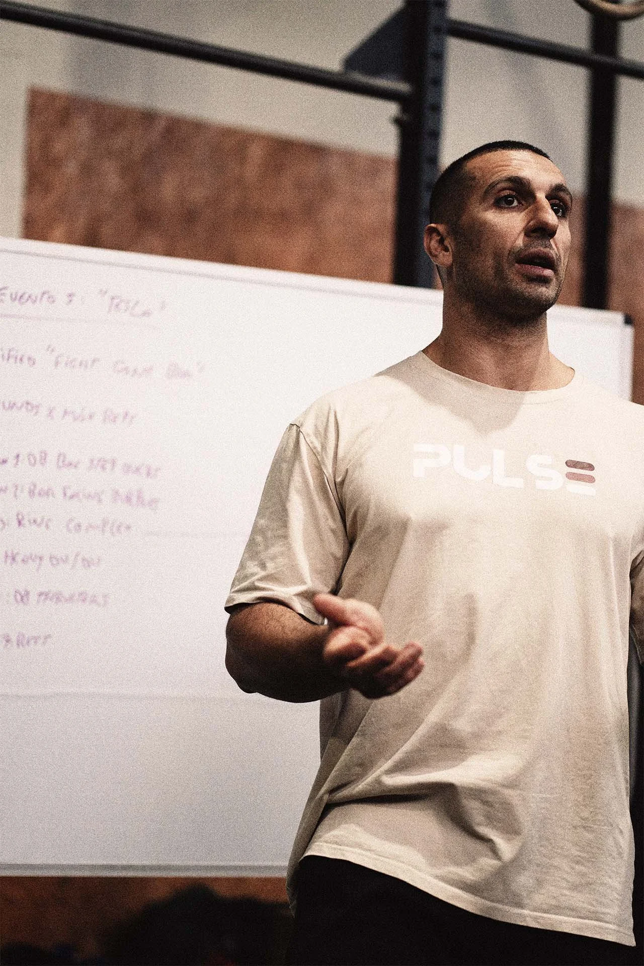 Un hombre dando una charla en un salón de clases, con una pizarra blanca al fondo que tiene texto escrito y lleva una camiseta beige con el logo "PULSE" en el frente.