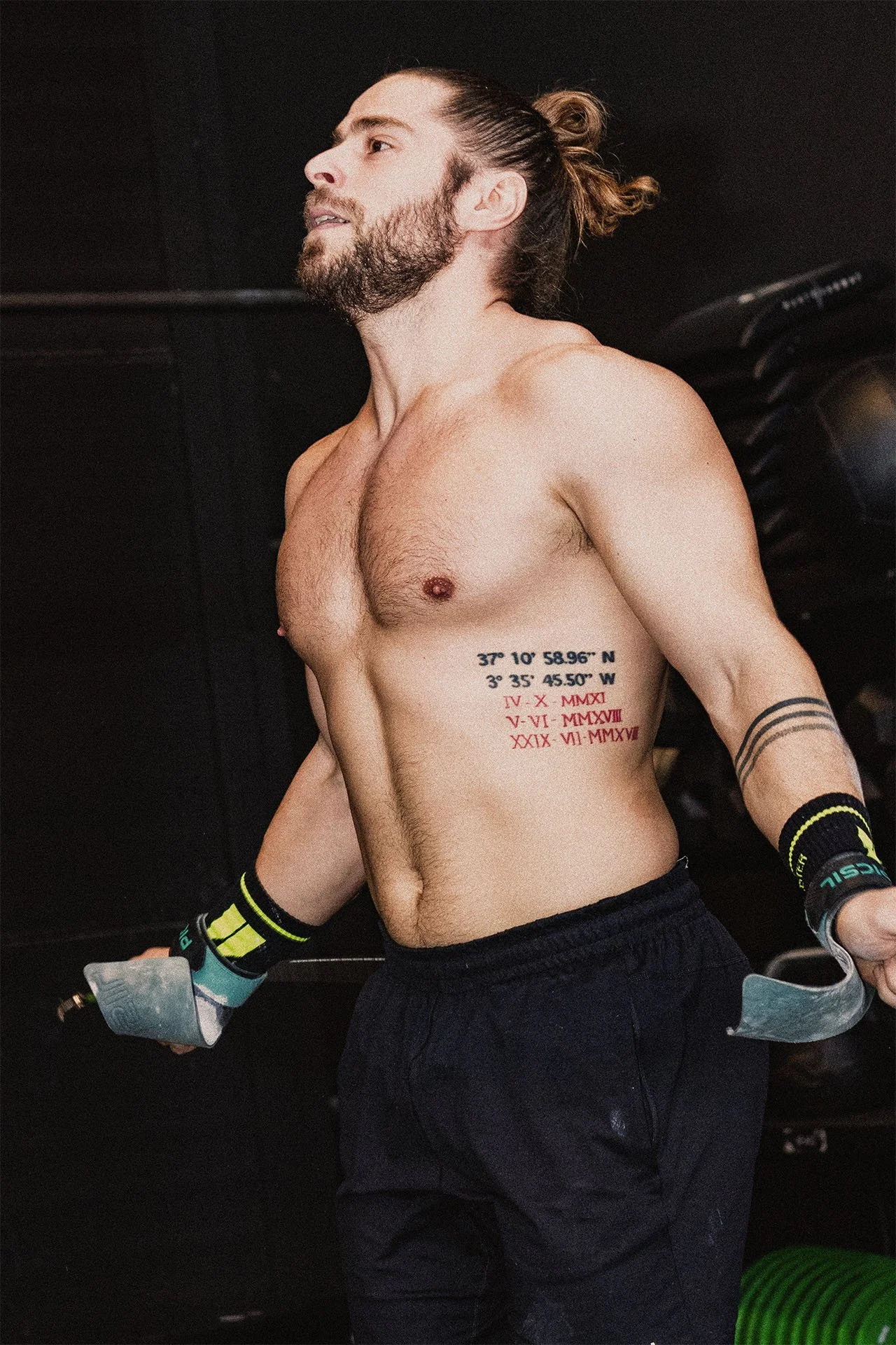 Hombre sin camiseta con tatuajes en el costado y el brazo, sosteniendo mancuernas en un gimnasio.