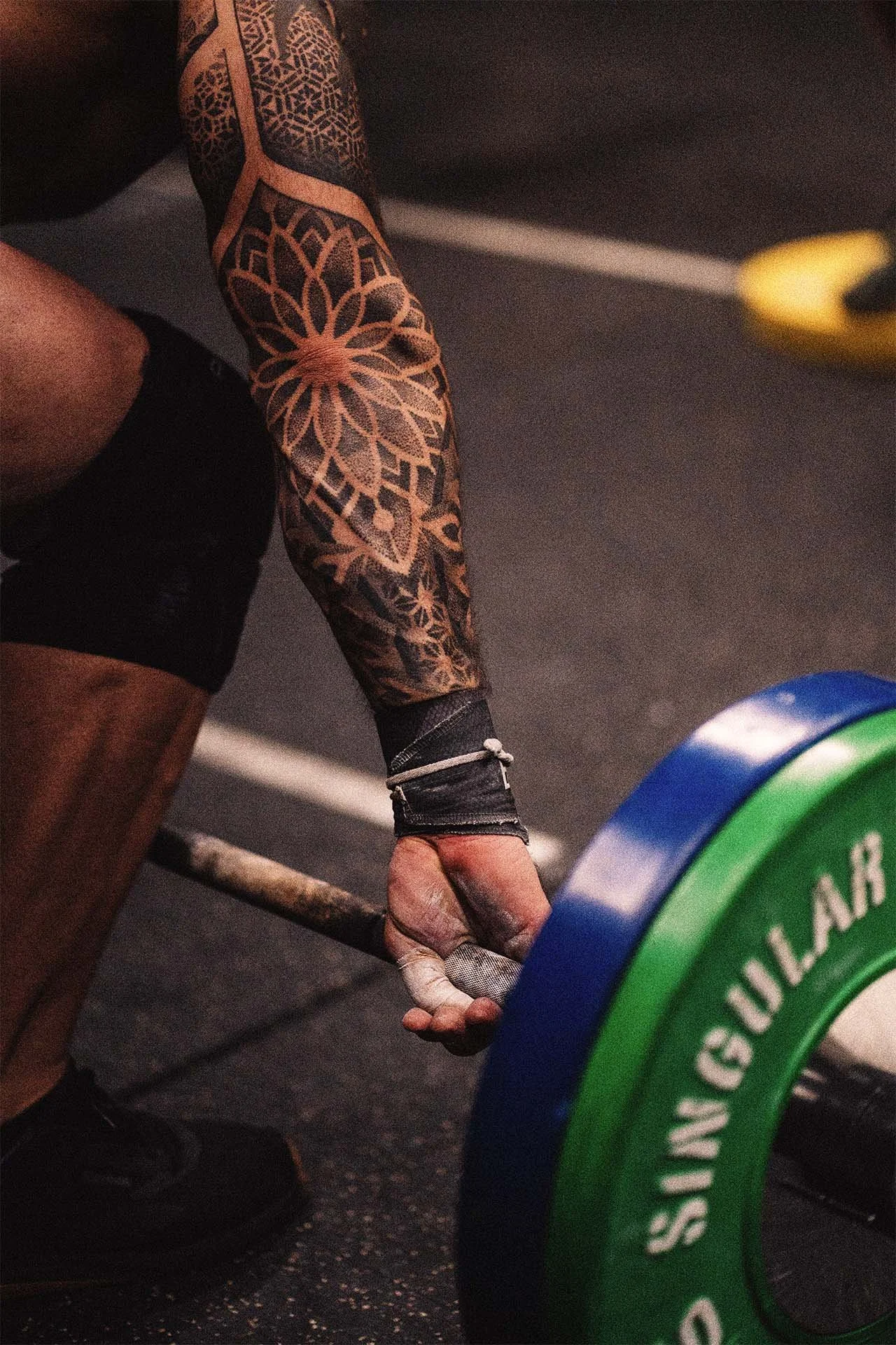 Persona levantando una barra con pesas en un gimnasio, tiene un tatuaje en el brazo, pantalones cortos negros y vendaje en la mano derecha.