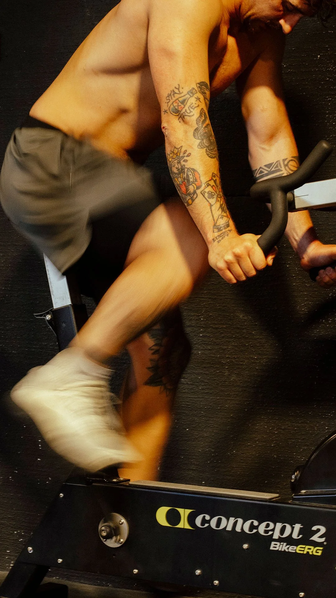 Hombre con tatuajes, sin camisa, usando pantalones cortos y zapatos blancos, montado en un ciclómetro en un gimnasio, haciendo ejercicio.
