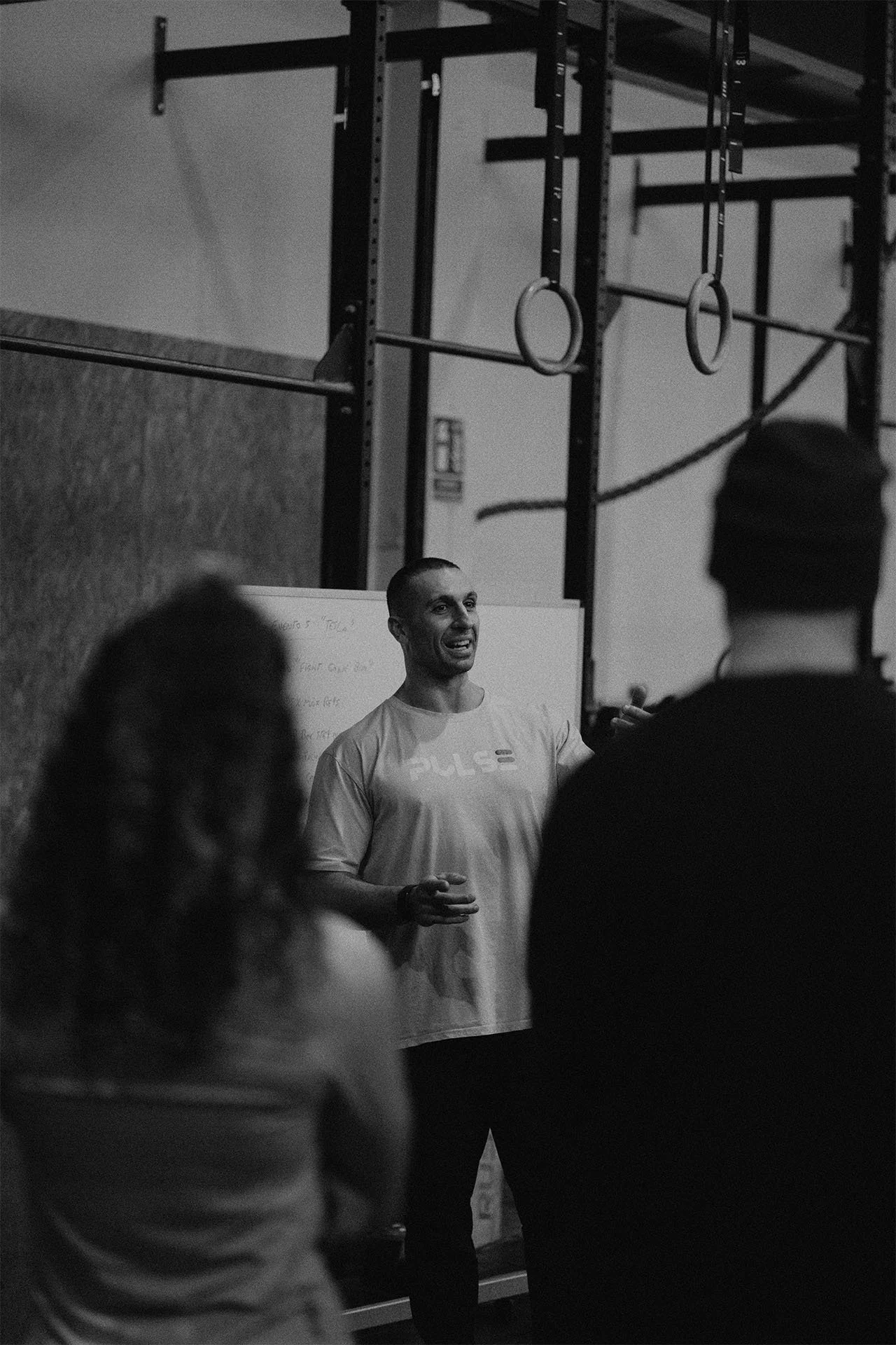 Un entrenador da una explicación a un grupo de personas en un gimnasio, con equipo de ejercicio en el fondo.