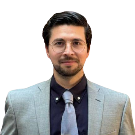 A man in a light gray suit with dark hair, beard, and glasses, standing against a bright background.