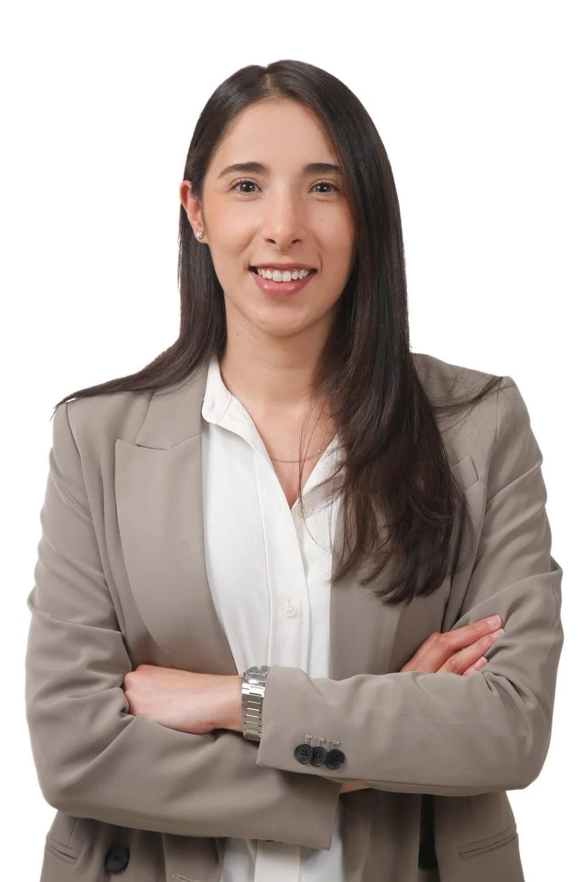 A woman with long dark hair, wearing a beige blazer and white shirt, smiling and crossing her arms, against a white background.