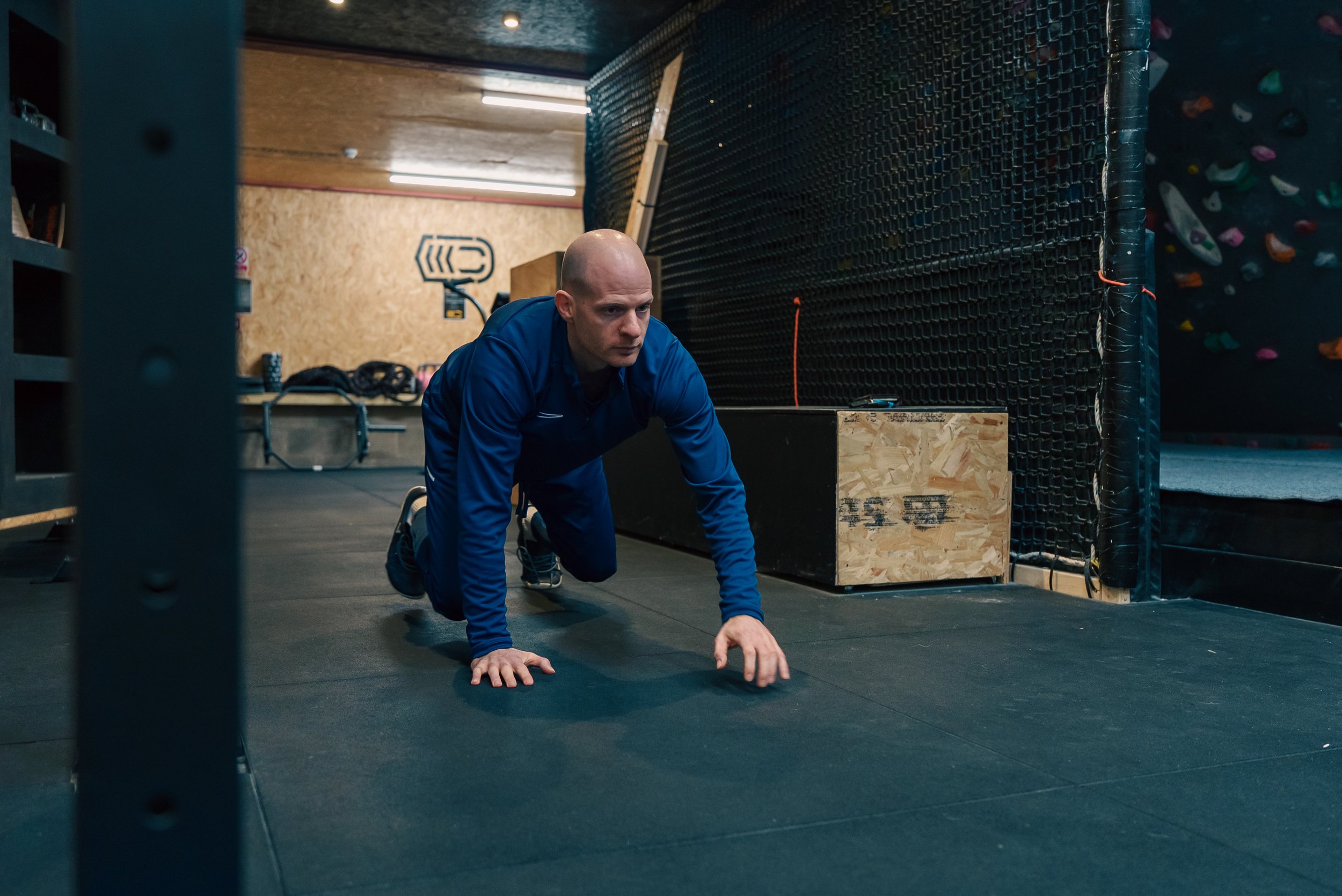 A man in blue athletic wear doing a mountain climber exercise on a black gym floor in a gym with climbing walls.