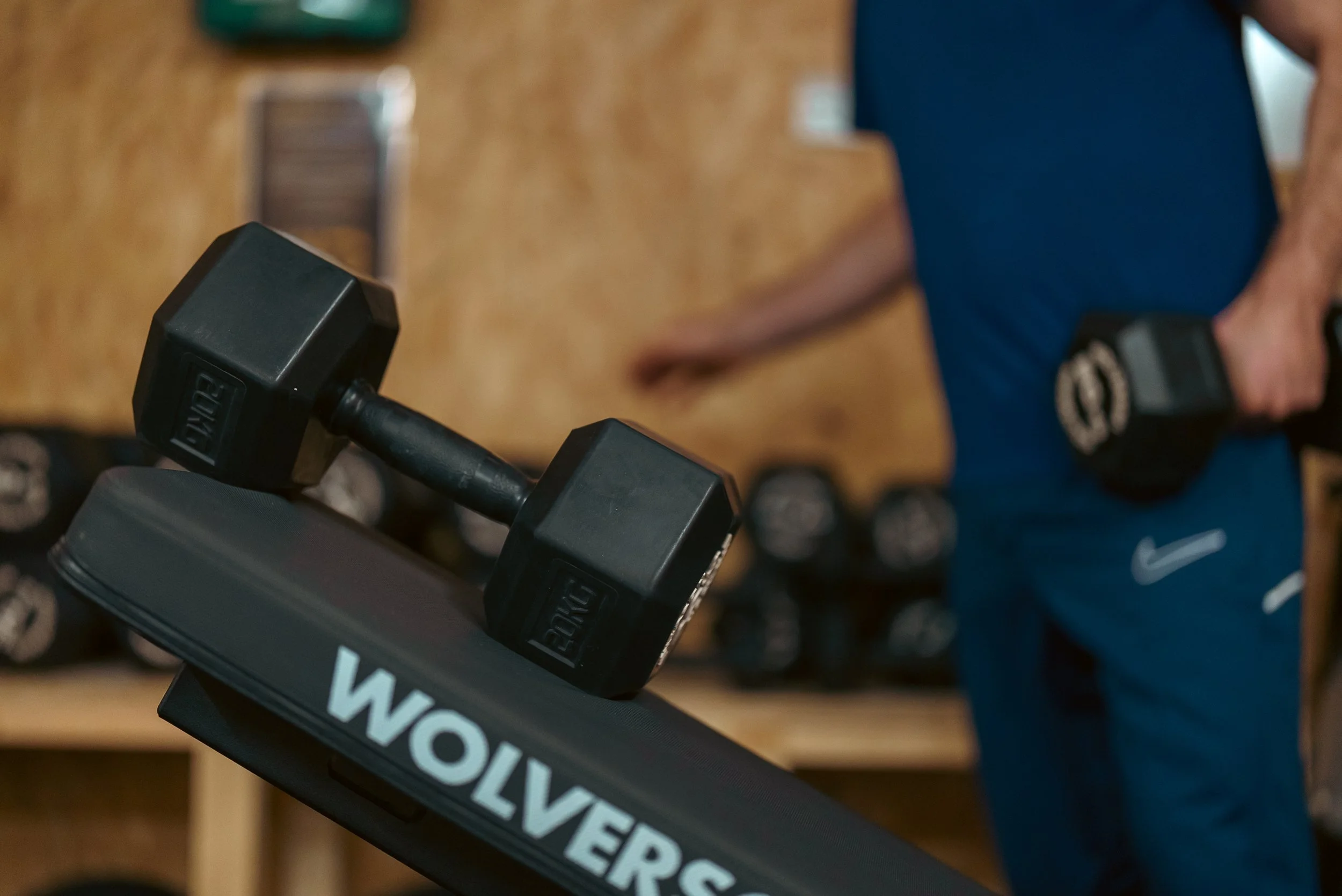 Close-up of a treadmill with the brand name 'WOLVER' visible, in a gym, with a person holding a dumbbell in the background.