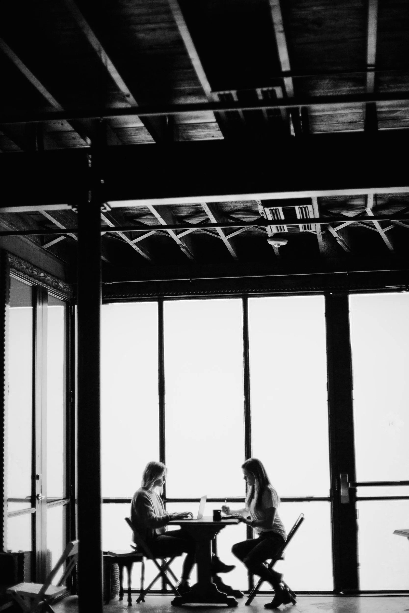 Two people sitting at a table in a modern room with large windows and a high ceiling. They are looking at their laptops, possibly working or having a discussion.