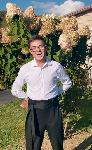 A young man with glasses smiling outdoors in front of large blooming hydrangea bushes, wearing a white button-up shirt and black pants.