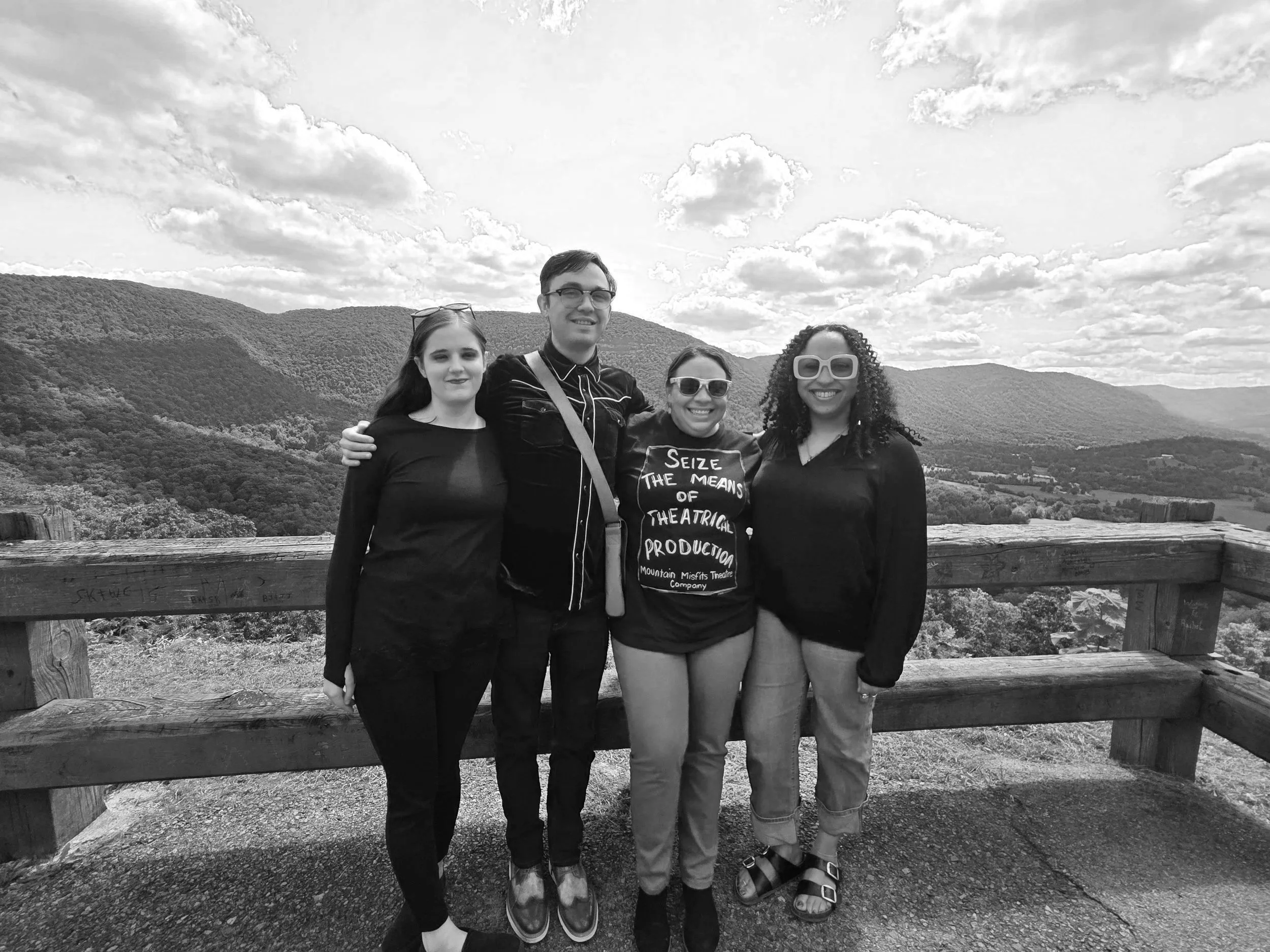 Four smiling people standing close together outdoors with a scenic mountain and cloudy sky in the background. One woman is wearing a t-shirt that reads 'Seize the means of theatrical production.'