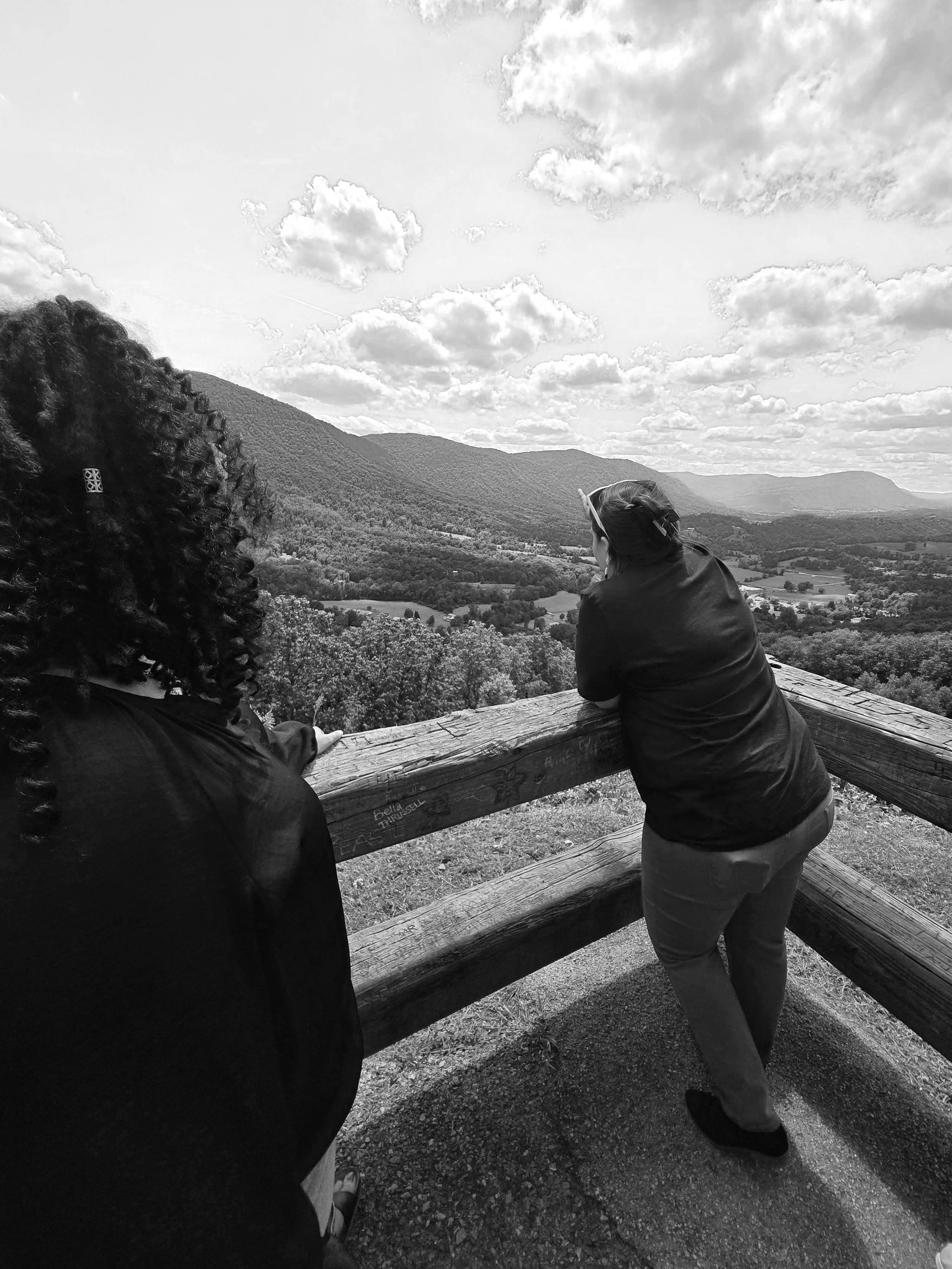 Two women standing at a wooden fence, overlooking a scenic view of rolling hills and mountains under a partly cloudy sky.