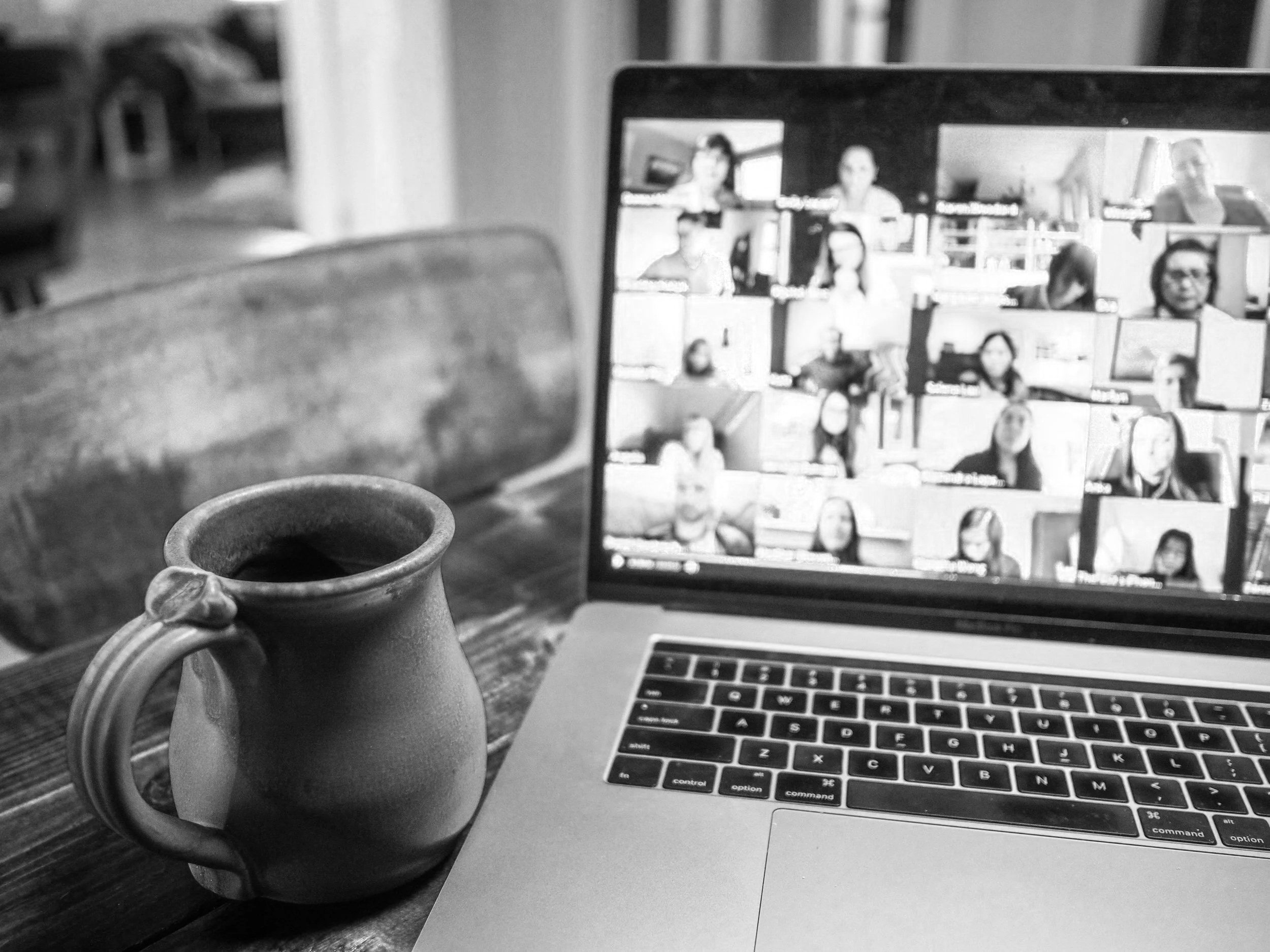A laptop on a wooden table, with a coffee mug in the foreground and a collage of people on the laptop screen, in a black-and-white photo.