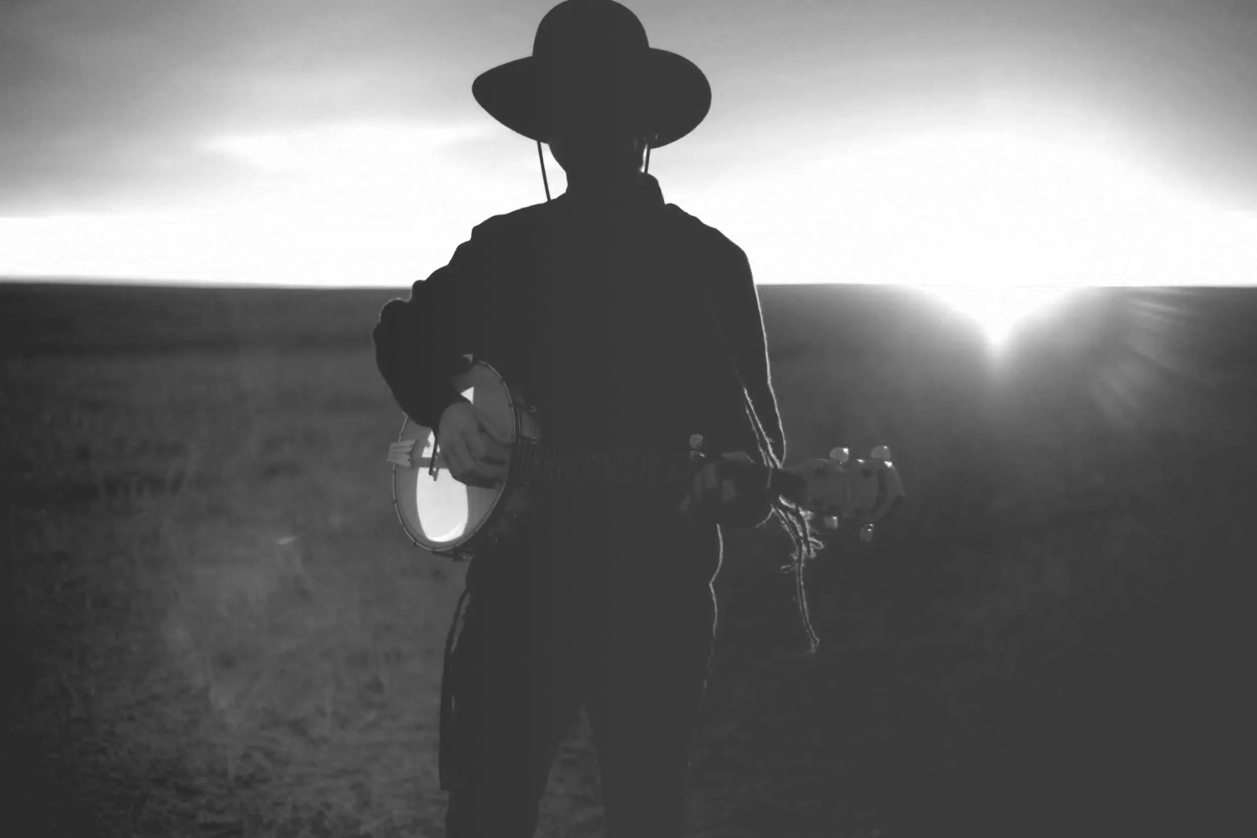 Silhouette of a person wearing a wide-brimmed hat and holding a guitar, standing outdoors near a body of water with a bright setting sun in the background.