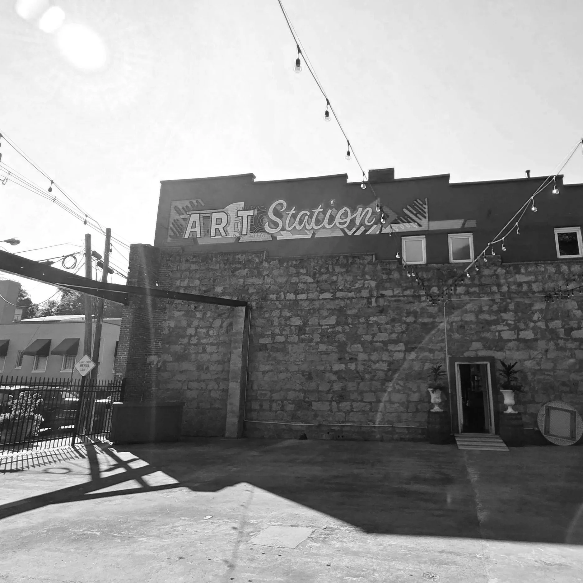 Street view of an art studio building with a brick wall, string lights overhead, a gated entrance on the left, and a small open gate on the right. The sky is clear with strong shadows on the ground.