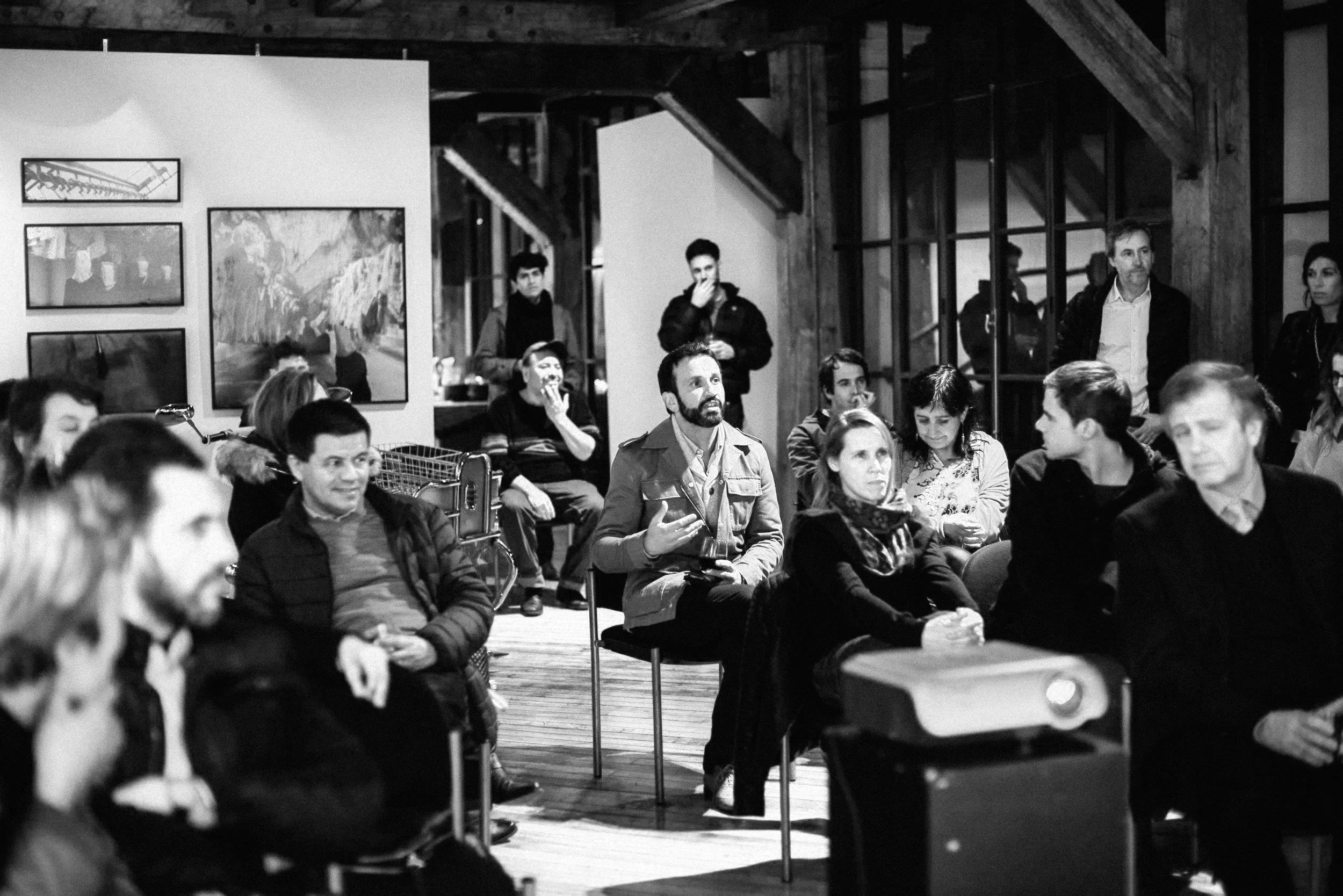 Black and white photo of a group of people seated indoors, possibly at an event or gathering. Several framed pictures hang on the wall, and some individuals are engaged in conversation or looking around, with one person in the center wearing a jacket and gesturing with their hands.