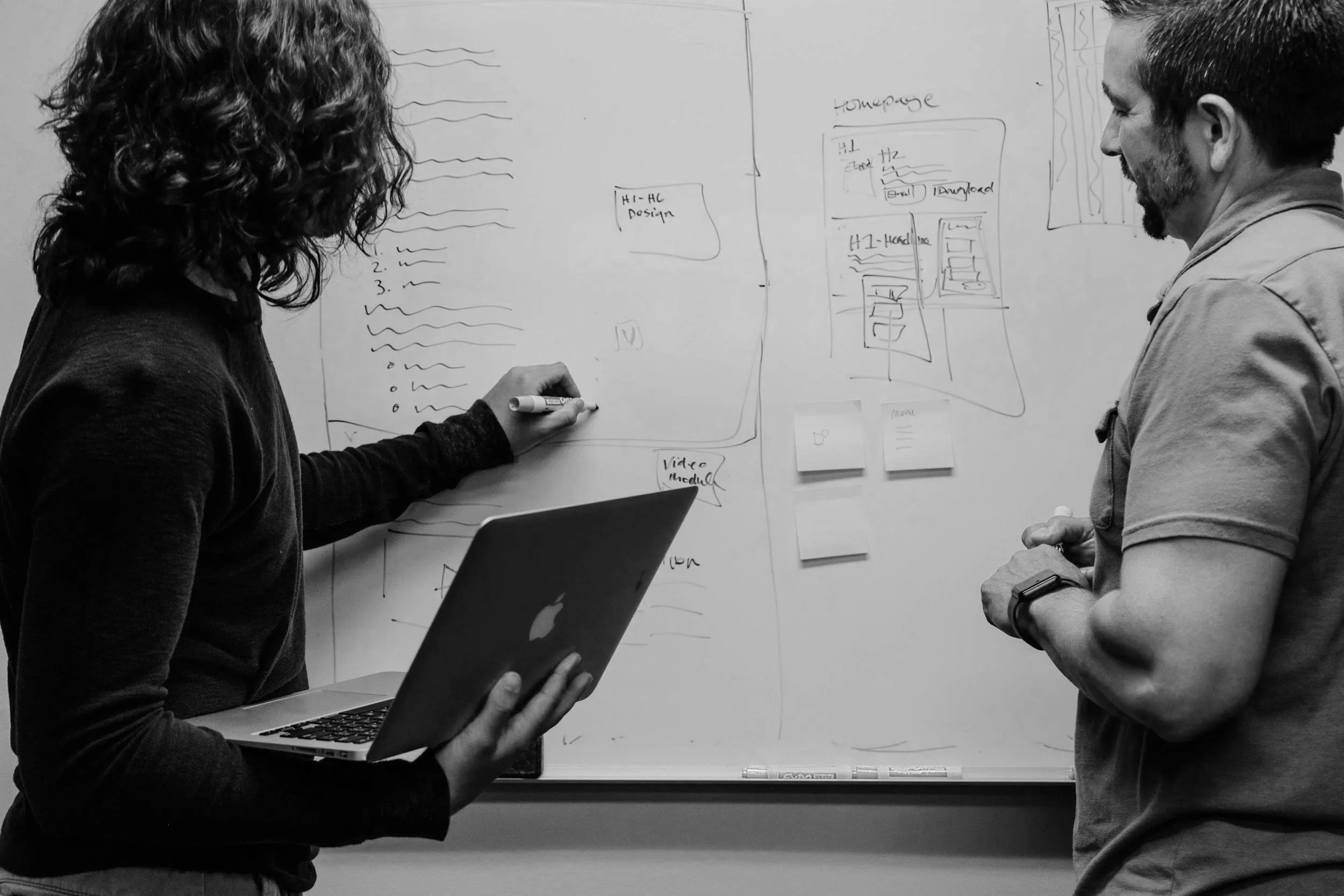 Two people standing and discussing in front of a whiteboard with sketches and notes, one holding a laptop and a marker.