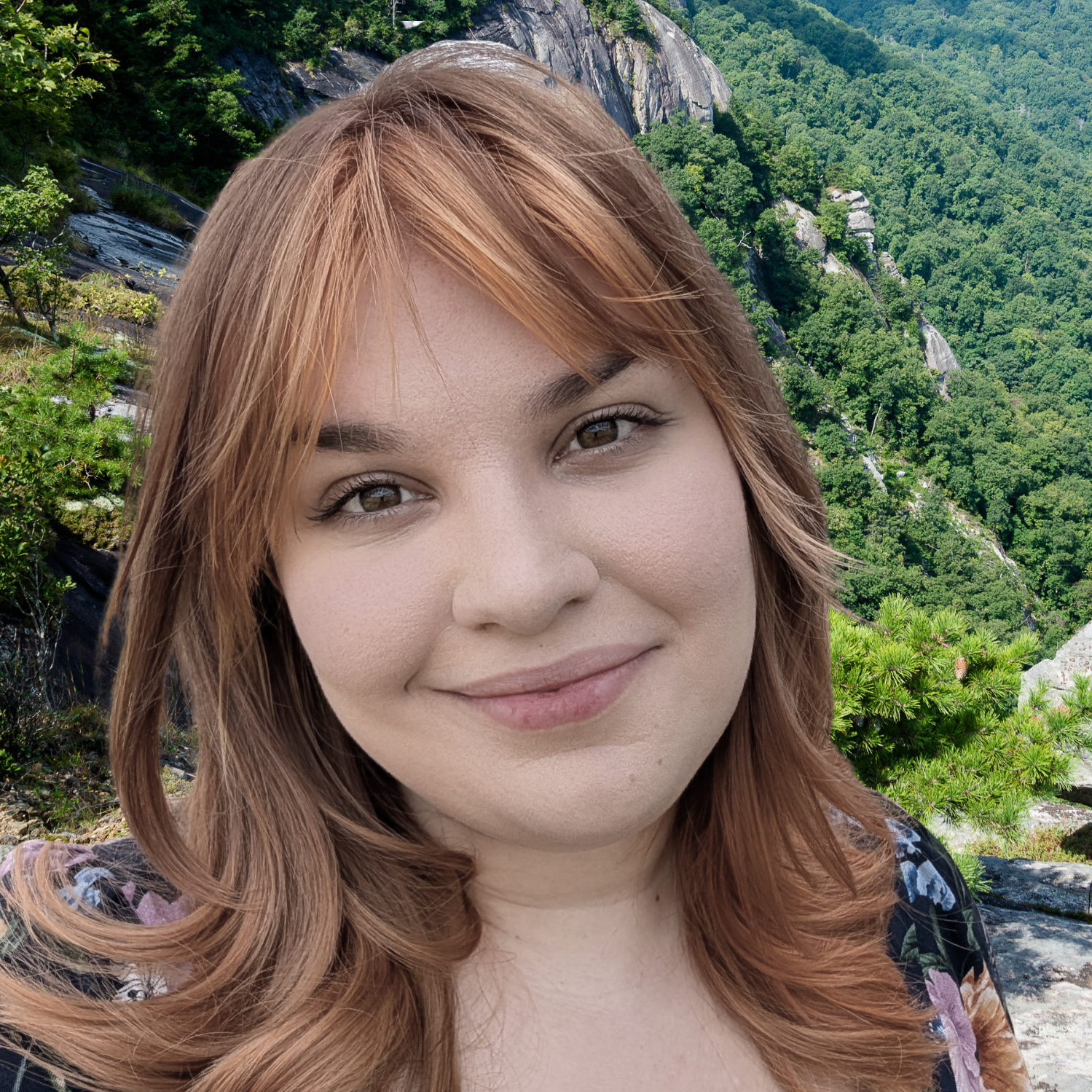 Close-up of a smiling woman with reddish-brown hair, in front of a lush green forest and rocky mountains.