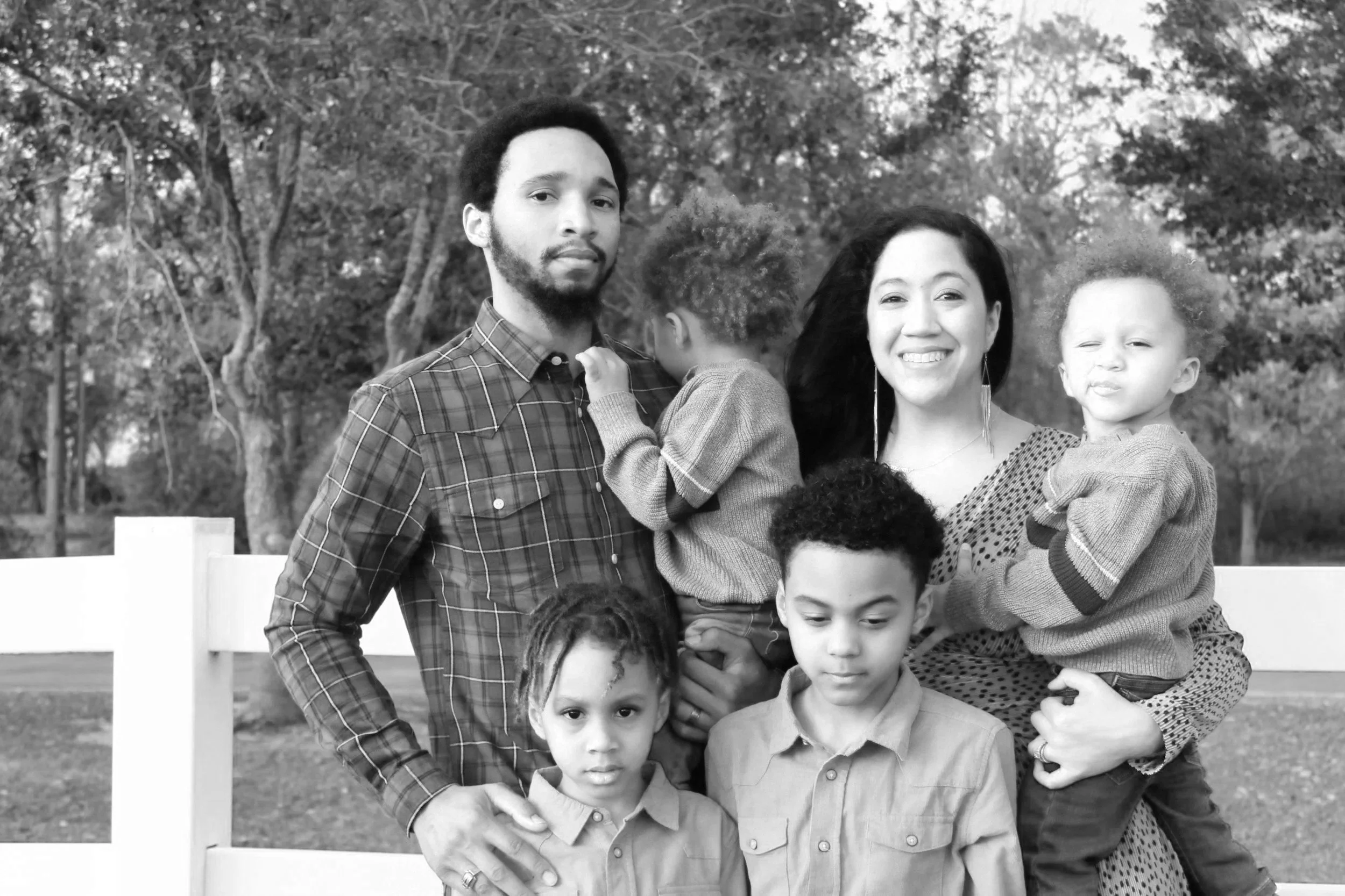 A family of six people outdoors in a park, including a mixed-race man with a beard, an Asian-looking woman with curly hair, and four children of various ages, standing in front of trees.