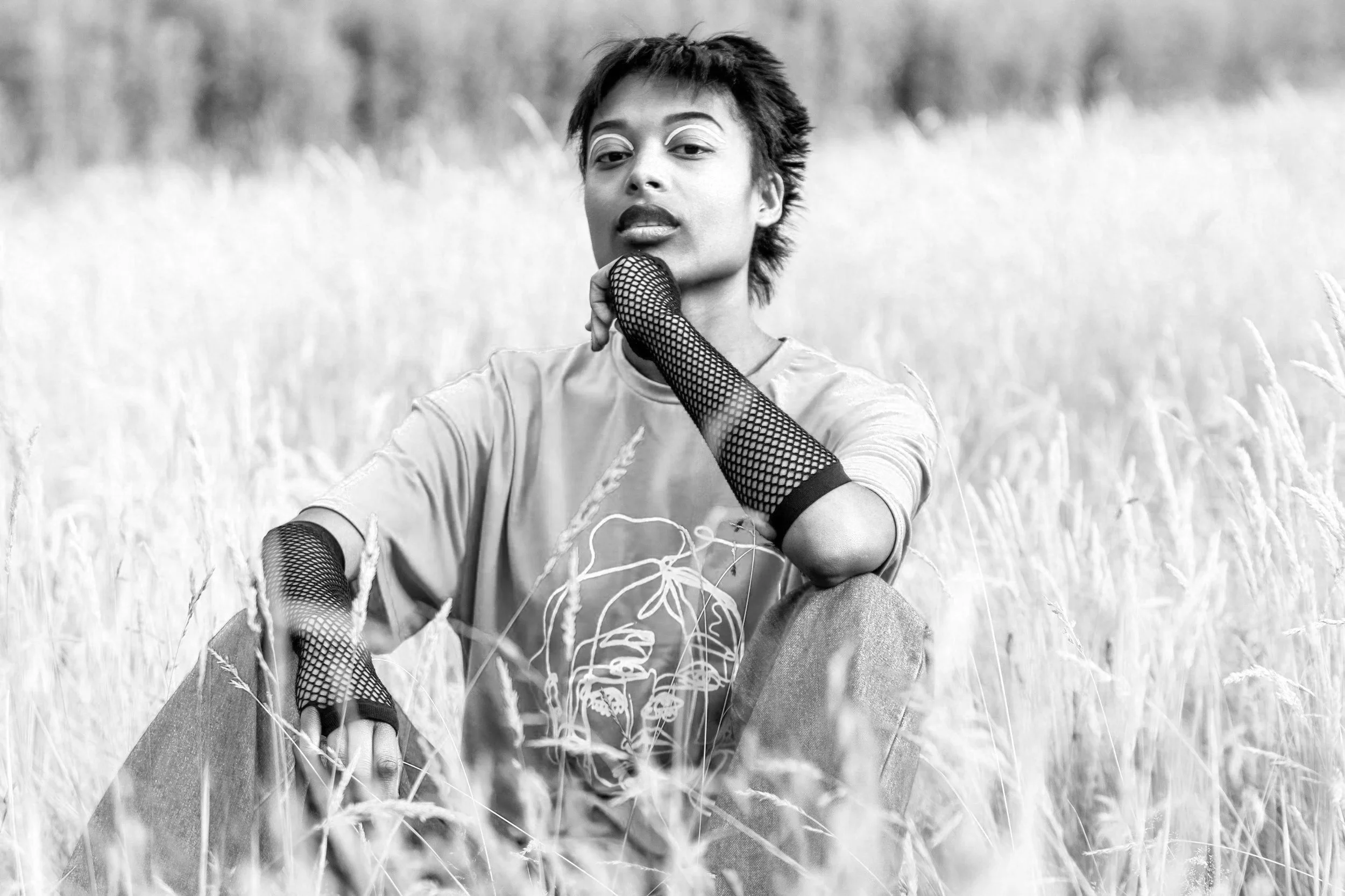 Black and white photo of a person with short hair, wearing fishnet gloves and a t-shirt, sitting in tall grass outdoors.