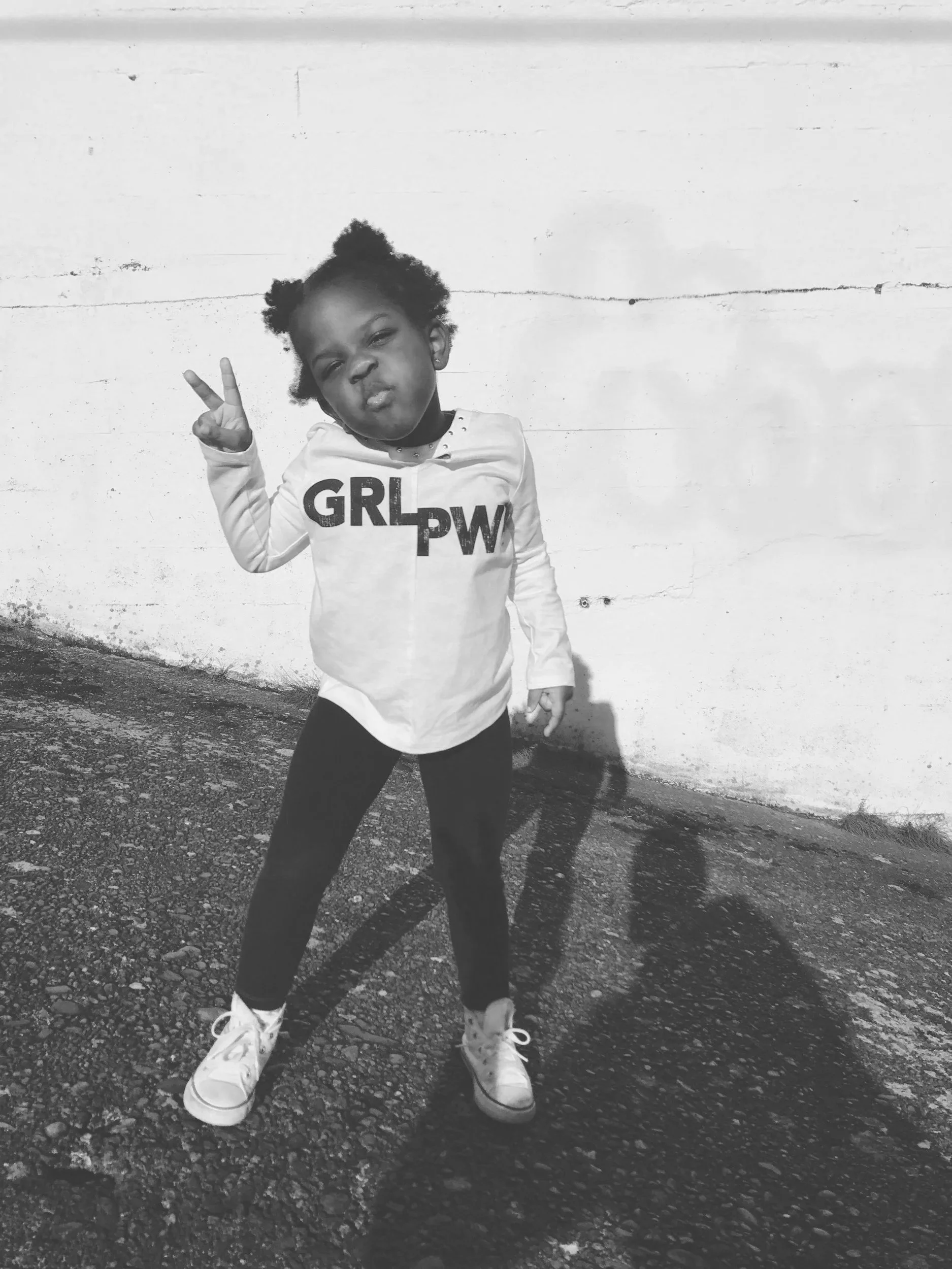 A young girl with puffy hair in pigtails making a peace sign with her right hand, standing against a white wooden wall. She has a playful, slightly serious expression, wearing a long-sleeved shirt with the words 'GRL PW' on the front, dark pants, and white sneakers.