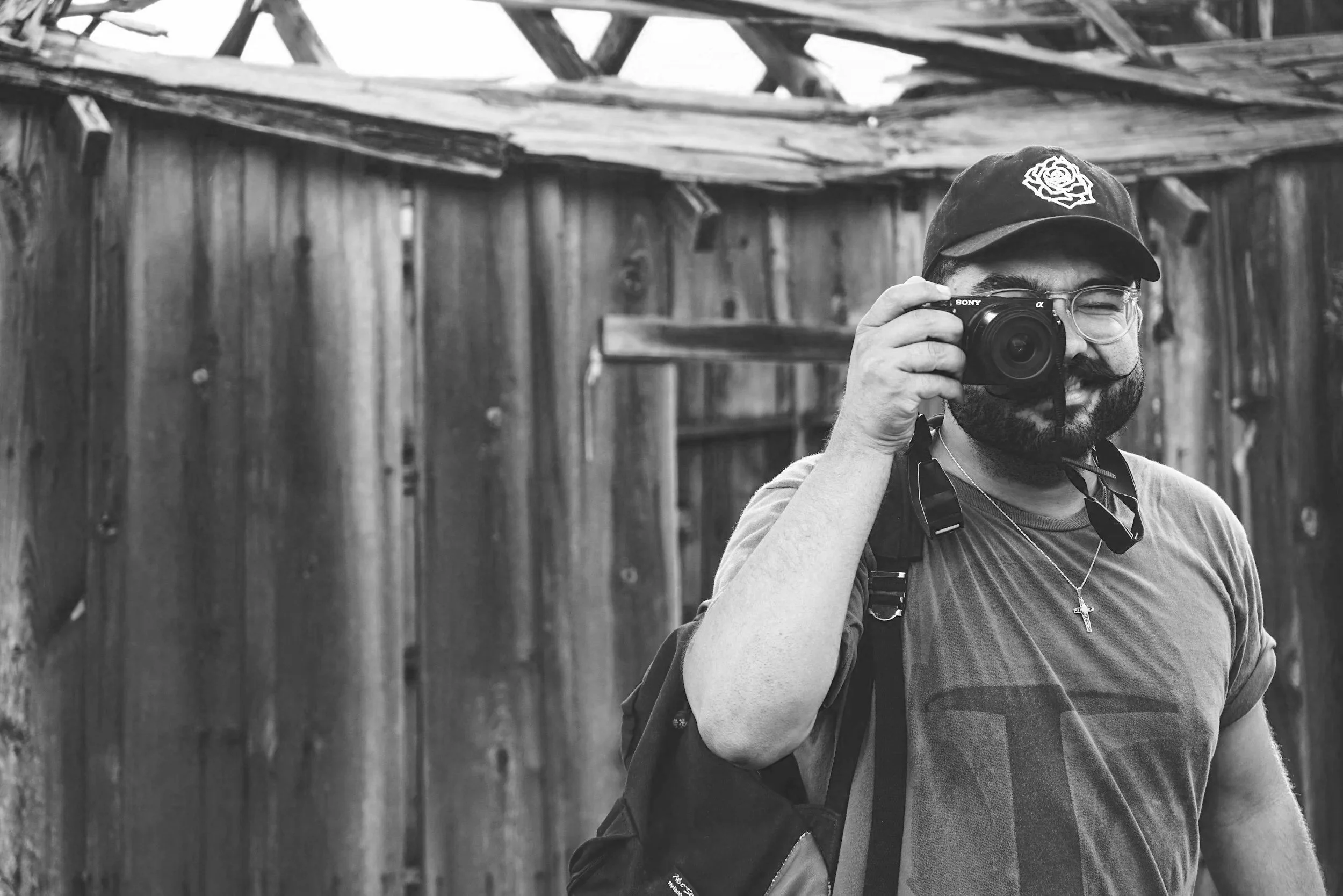 A man with glasses and a beard takes a photo with a Sony camera, standing in front of a wooden fence and barn in black and white.