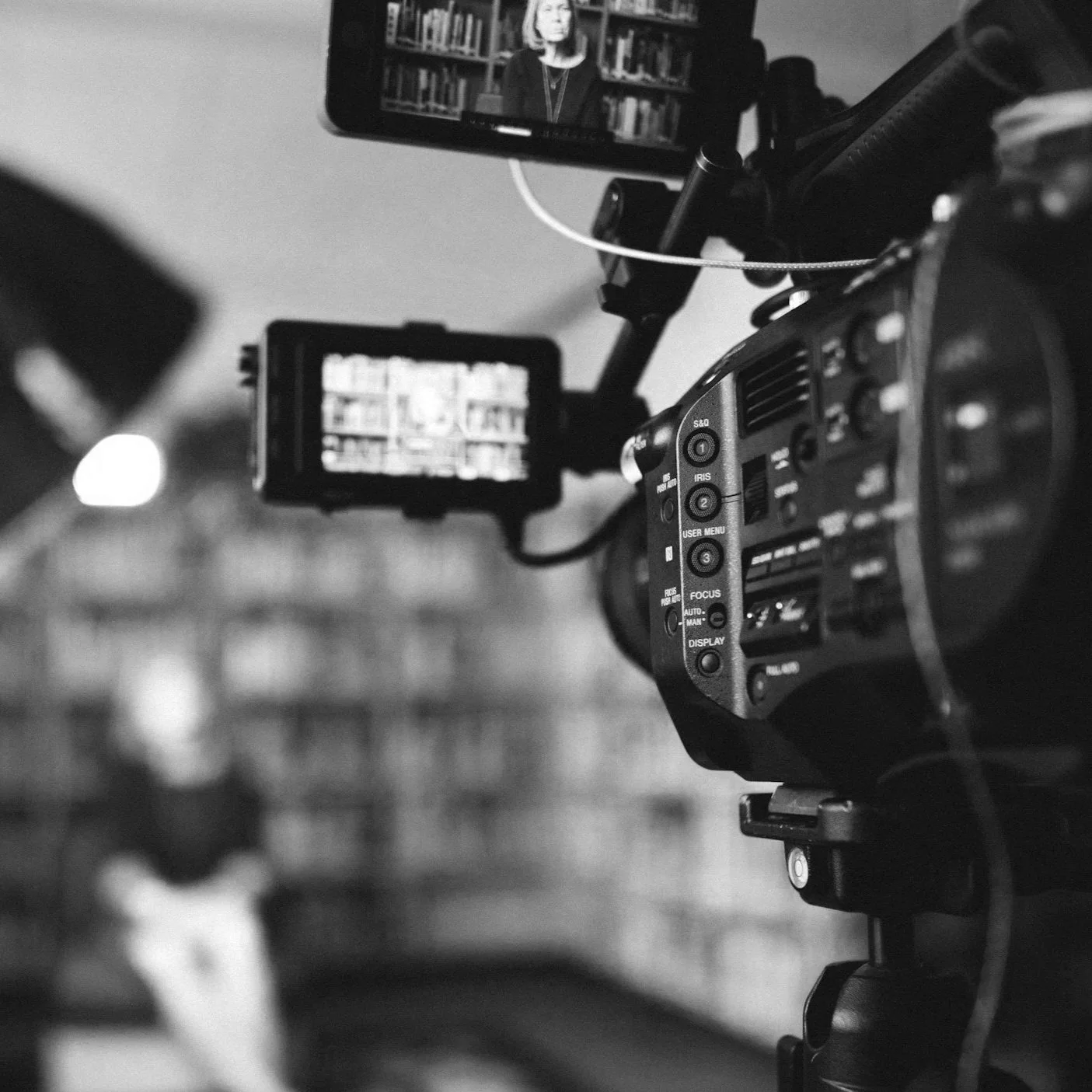 Black and white photo of a professional camera capturing a woman speaking in front of a bookshelf.