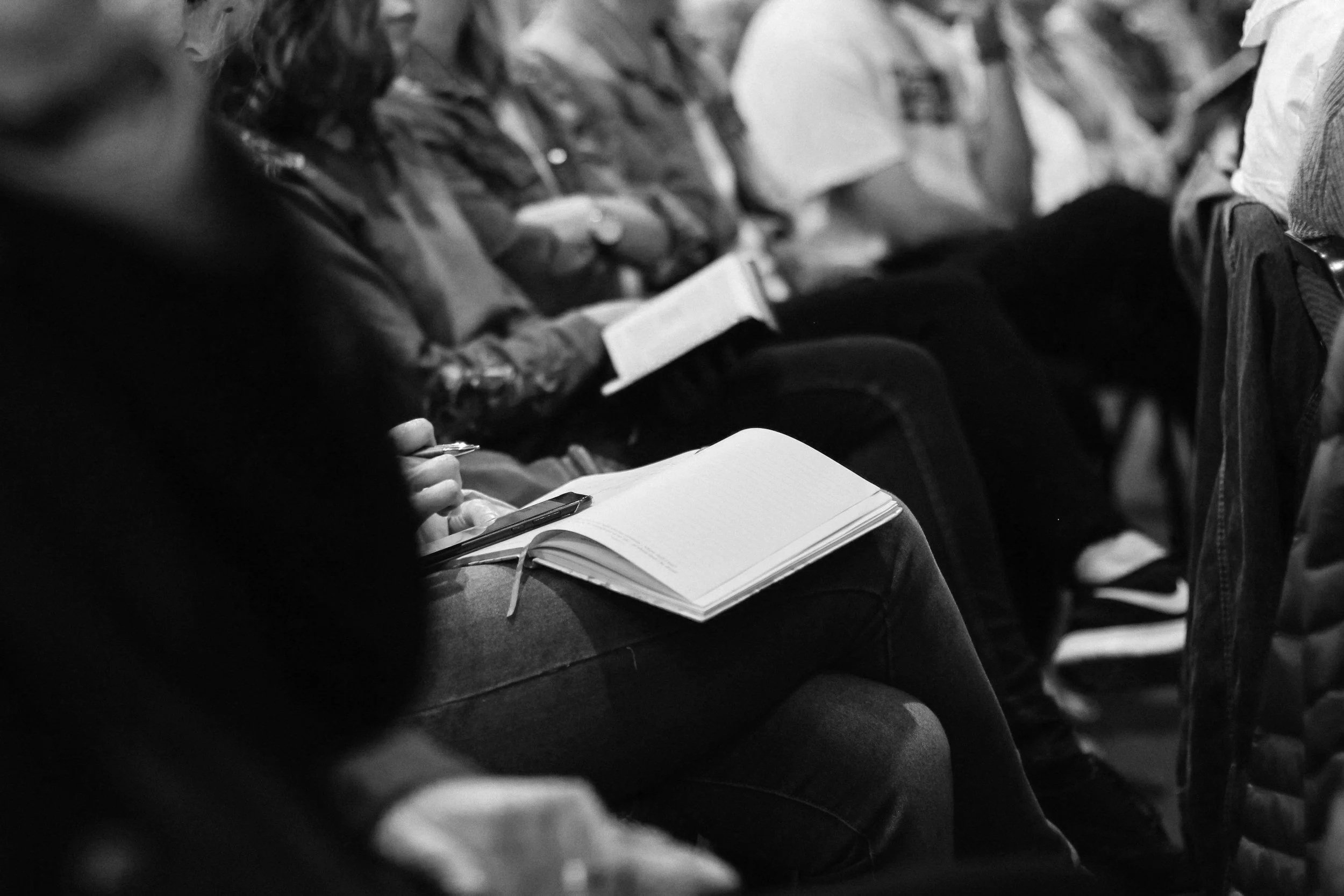 Black and white photo showing people sitting in a row, some holding notebooks or papers, attending a lecture or seminar.