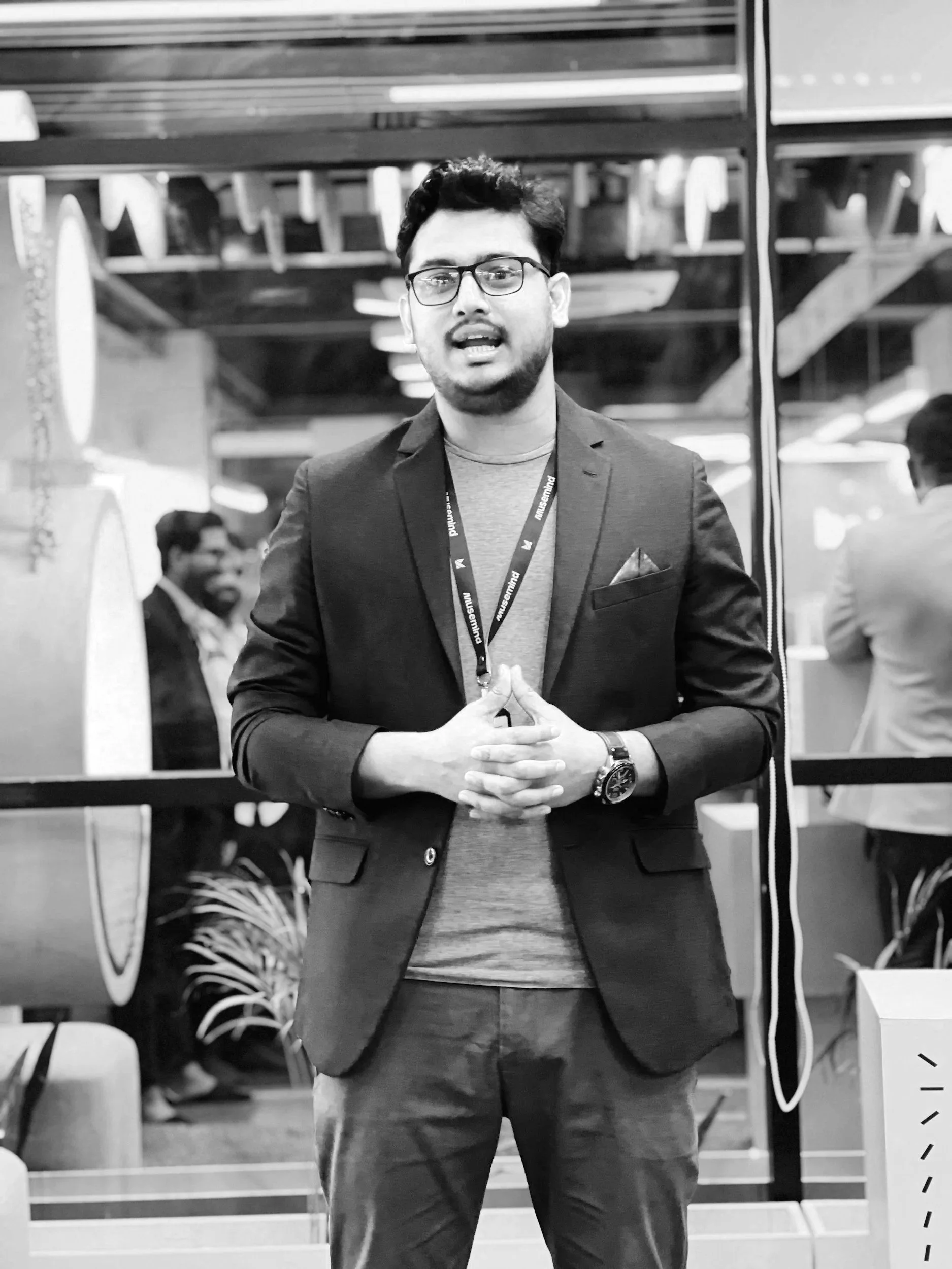 Black and white photo of a man wearing glasses, a suit jacket, and a lanyard, standing in an indoor setting with people blurred in the background.