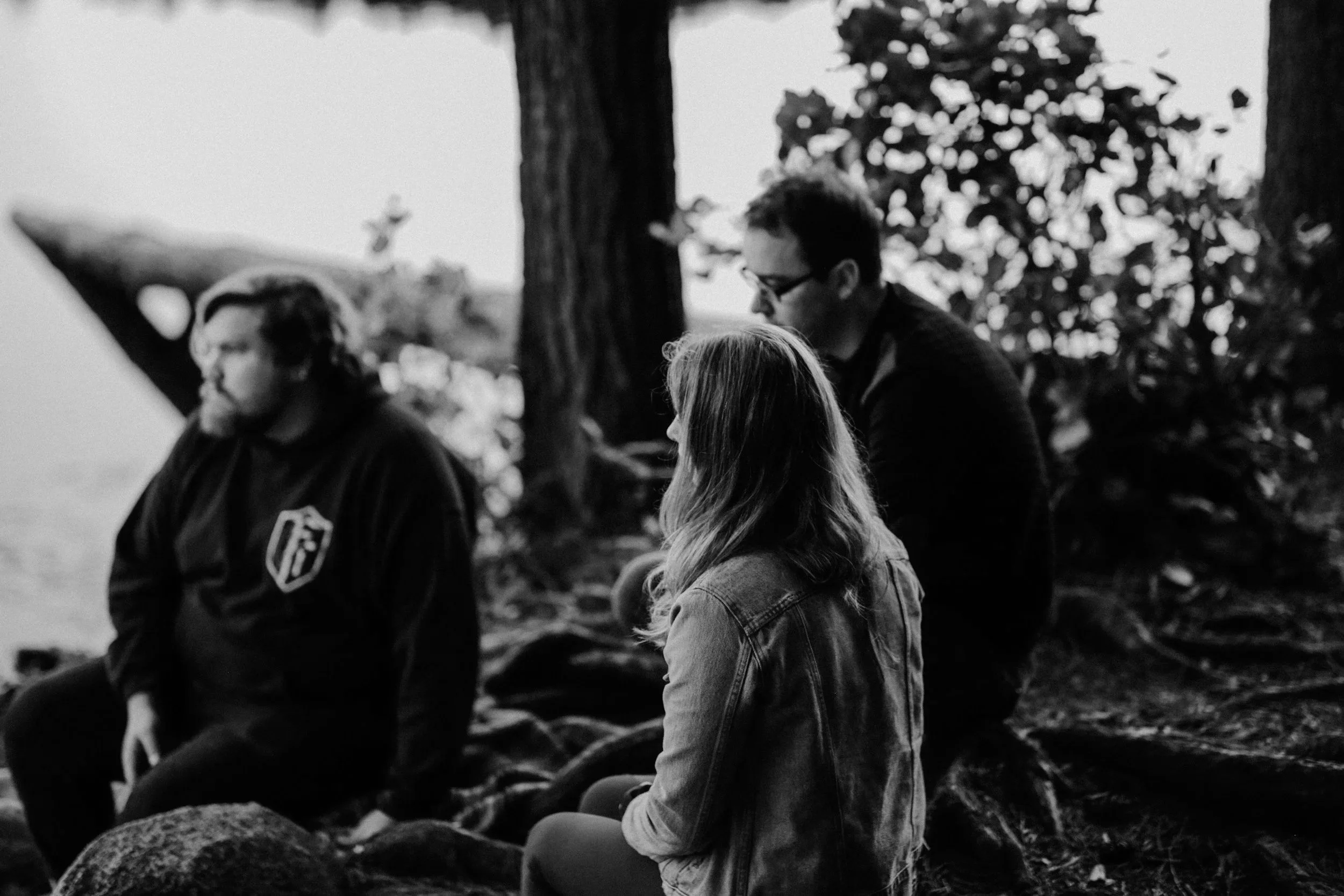 Three people sitting outdoors on rocks beneath trees, appears to be a candid black-and-white photograph of two women and one man, all with long hair, with the woman in the middle having light-colored hair and the woman on the left having glasses and dark hair.