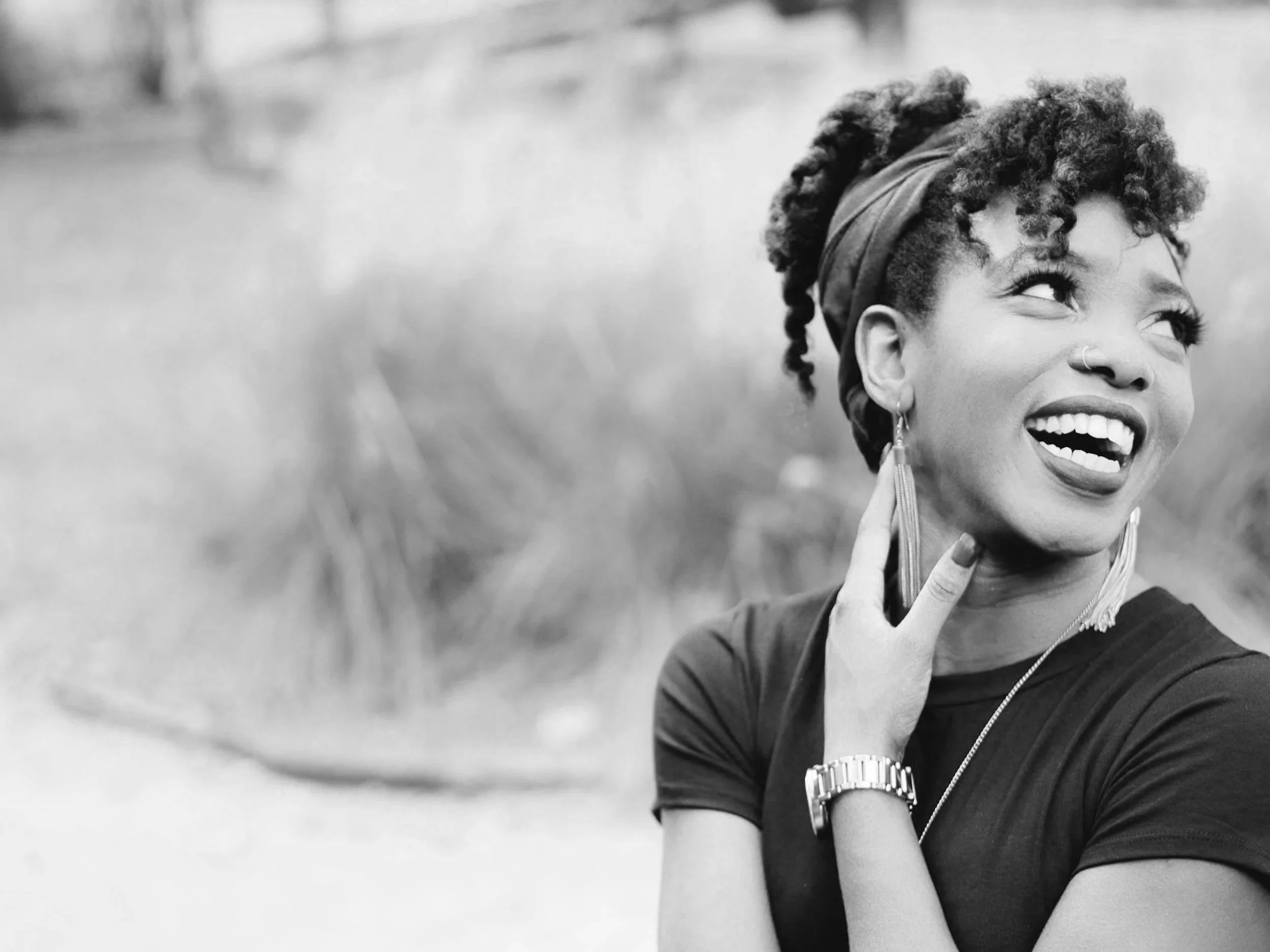 A smiling woman with curly hair tied back with a hairband, wearing earrings and a watch, looking up and to the right in an outdoor setting.