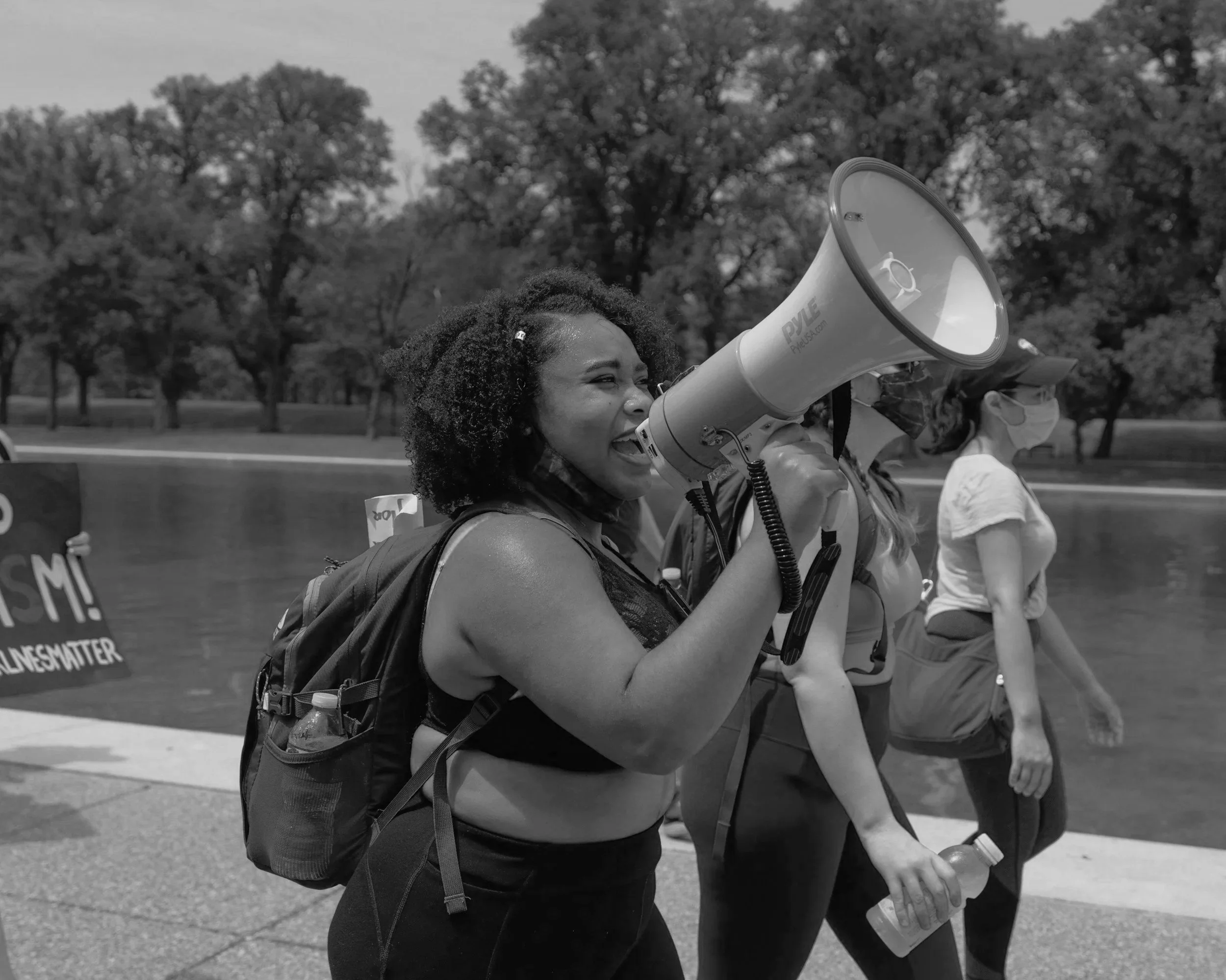 A woman with curly hair speaking into a megaphone during a protest or march, other participants visible in the background, outdoors with trees.