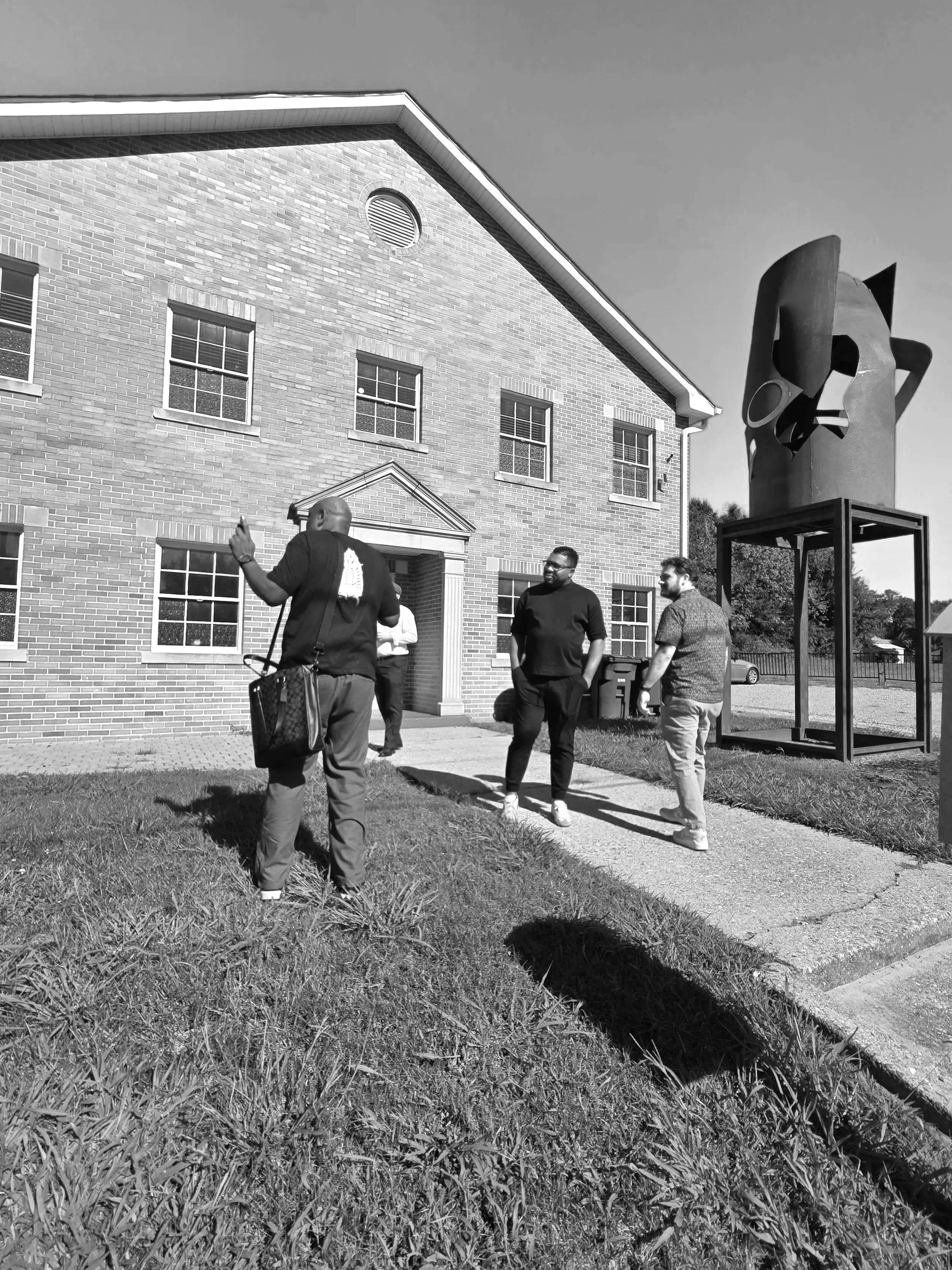 Four people standing and chatting outside a two-story brick building with multiple windows, with a large, abstract sculpture on a pedestal nearby, under a clear sky.