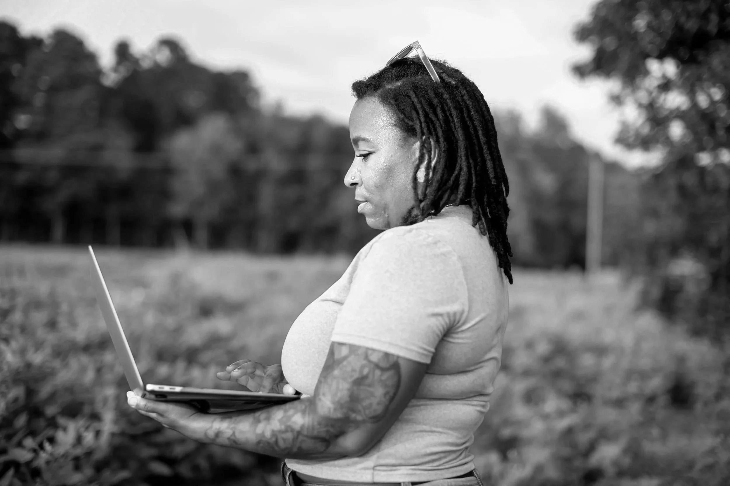 A woman with dreadlocks, sunglasses on her head, wearing a short-sleeved shirt, is looking at a laptop she is holding in her hands outdoors, with a blurred background of trees and sky.
