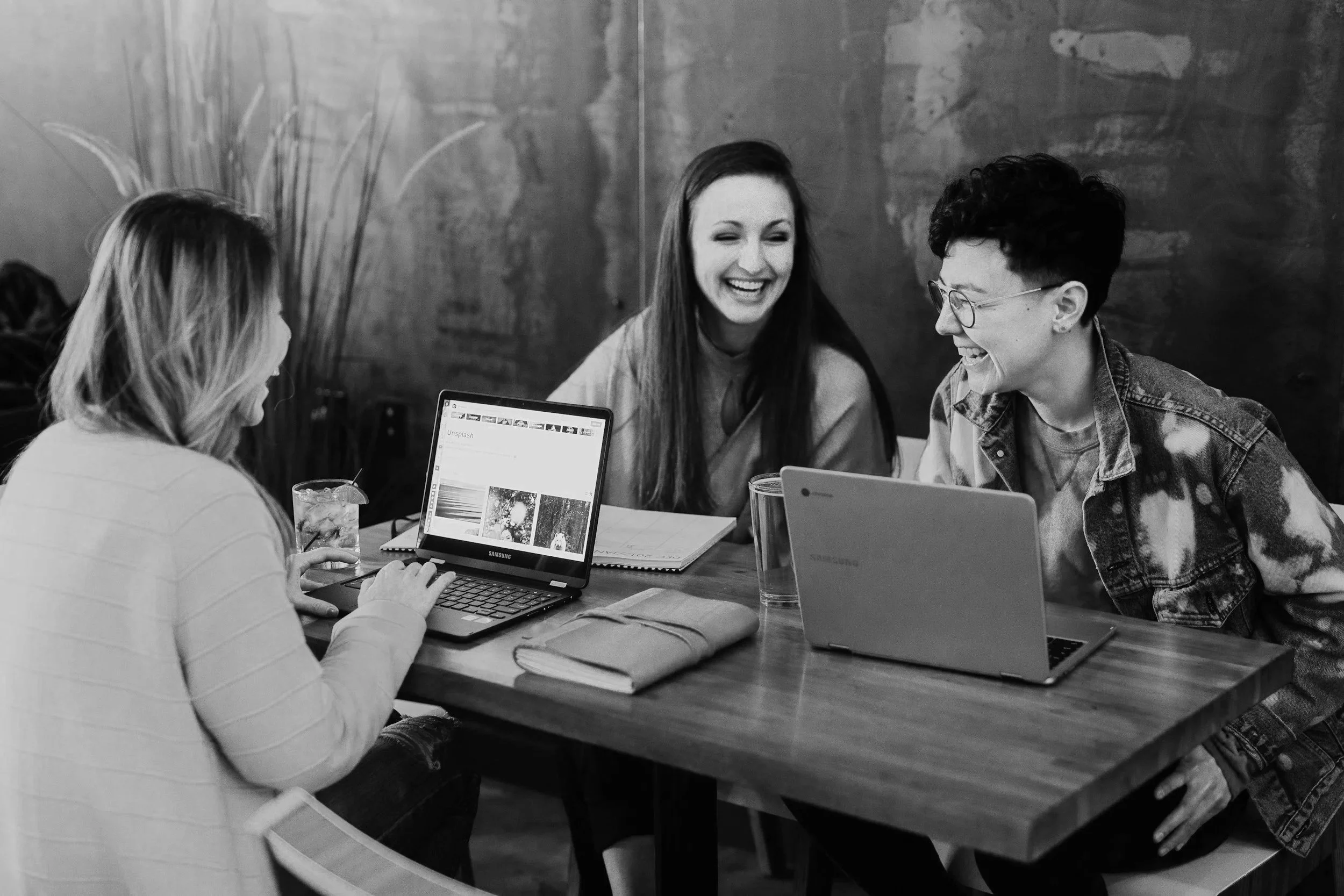Four young women sitting around a wooden table, engaging in conversation and laughing, with laptops and drinks in front of them in a casual indoor setting.