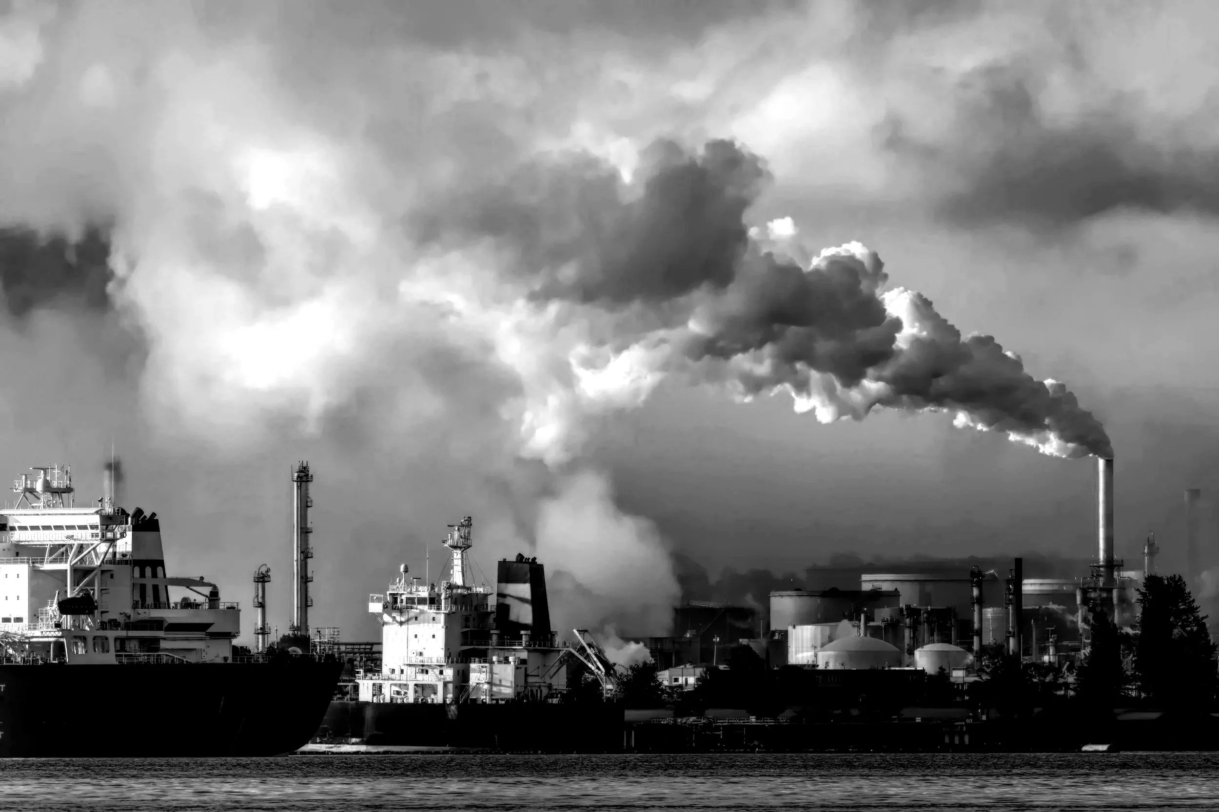 A black and white photo of an industrial port scene with multiple ships, smokestacks emitting pollution, and factory buildings near water.