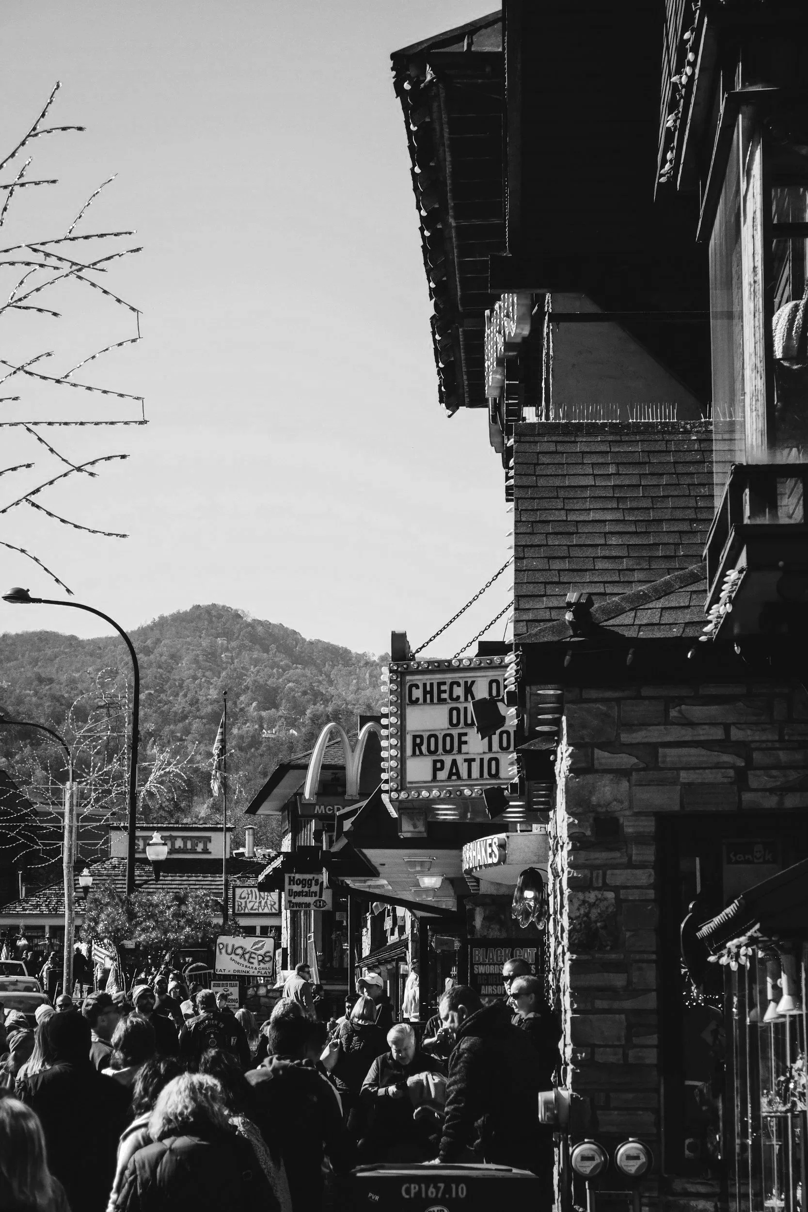 A busy street scene with a crowd of people, storefronts, and signage, including a 'CHECK O R OOF TO PATIO' sign, with rolling hills or mountains in the background.