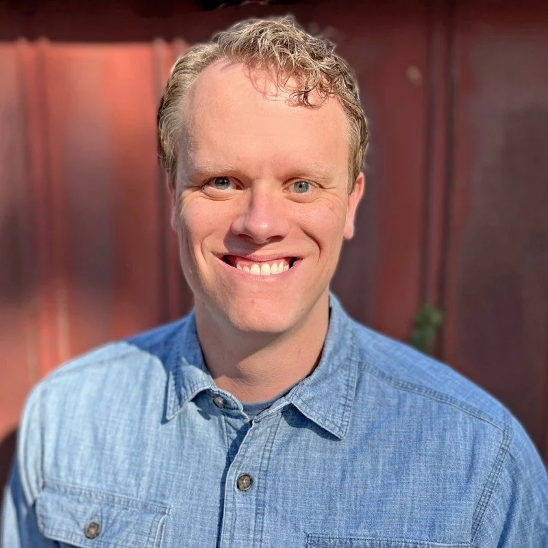 A man with short curly blonde hair and blue eyes smiling, wearing a light blue denim button-up shirt, standing outdoors with a blurred wooden fence background.
