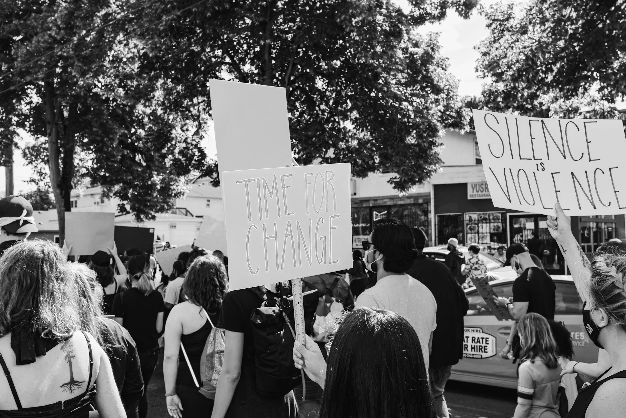 Crowd of people at a protest holding signs, one sign reads 'Time for Change' and another reads 'Silence is Violence,' with trees and buildings in the background, black and white photo.