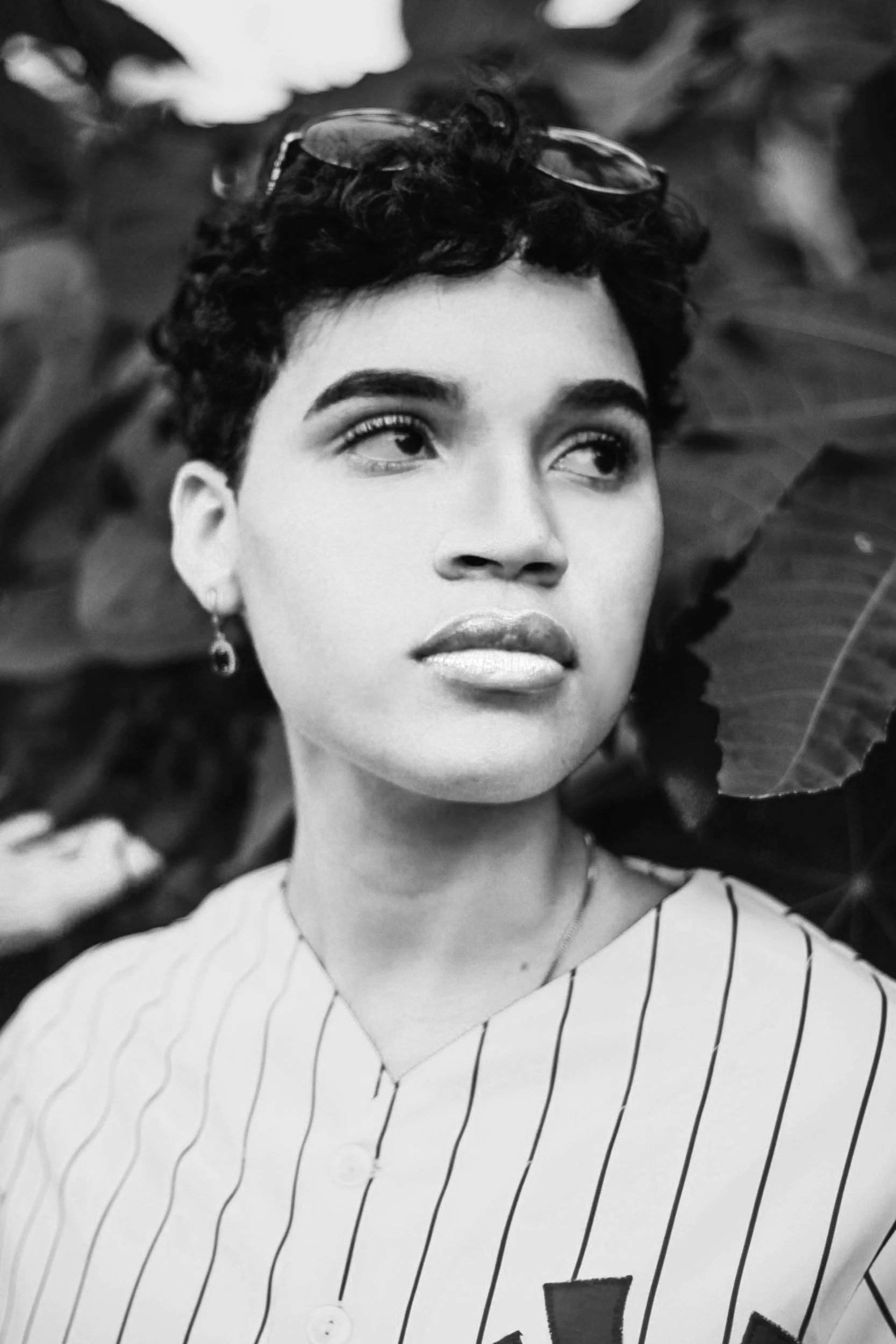 Close-up black and white photo of a young woman with short curly hair, wearing glasses on her head, a striped shirt, and an earring, surrounded by large leaves.