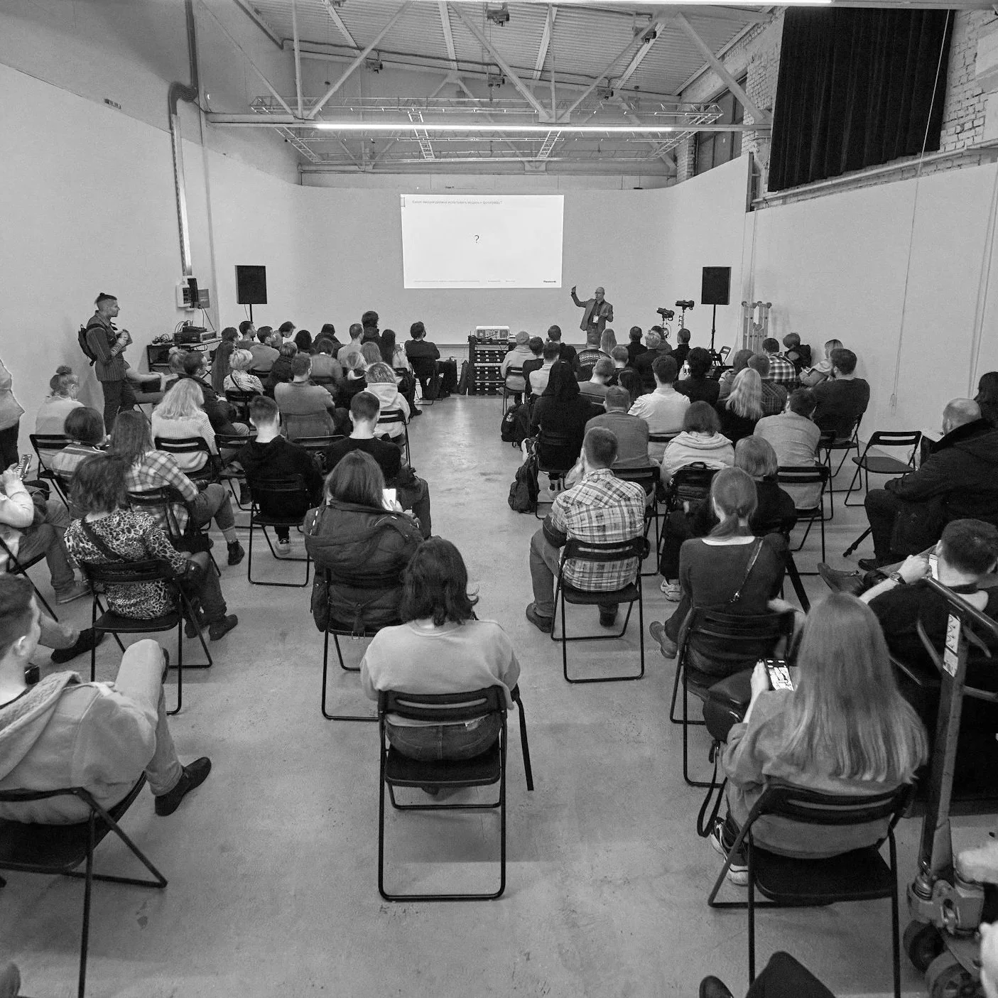 A large group of people is attending a presentation or lecture in a town hall with exposed brick walls and high ceilings. A speaker stands at the front near a large screen, gesturing with their hand. Some audience members are taking notes or photos.