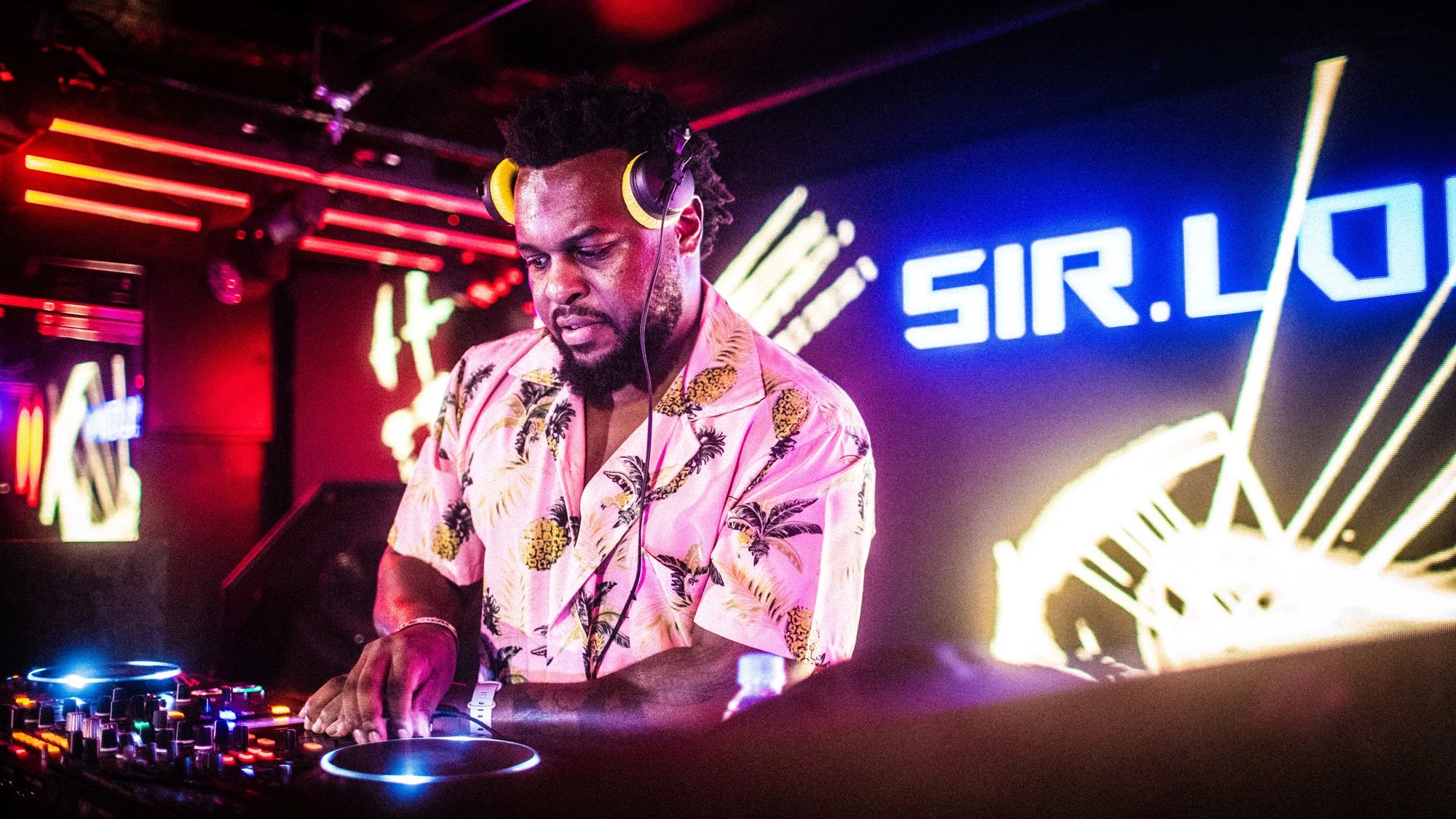 A DJ wearing headphones and a floral shirt is performing at a nightclub with colorful lighting and a neon sign that reads 'SIR.LO' in the background.