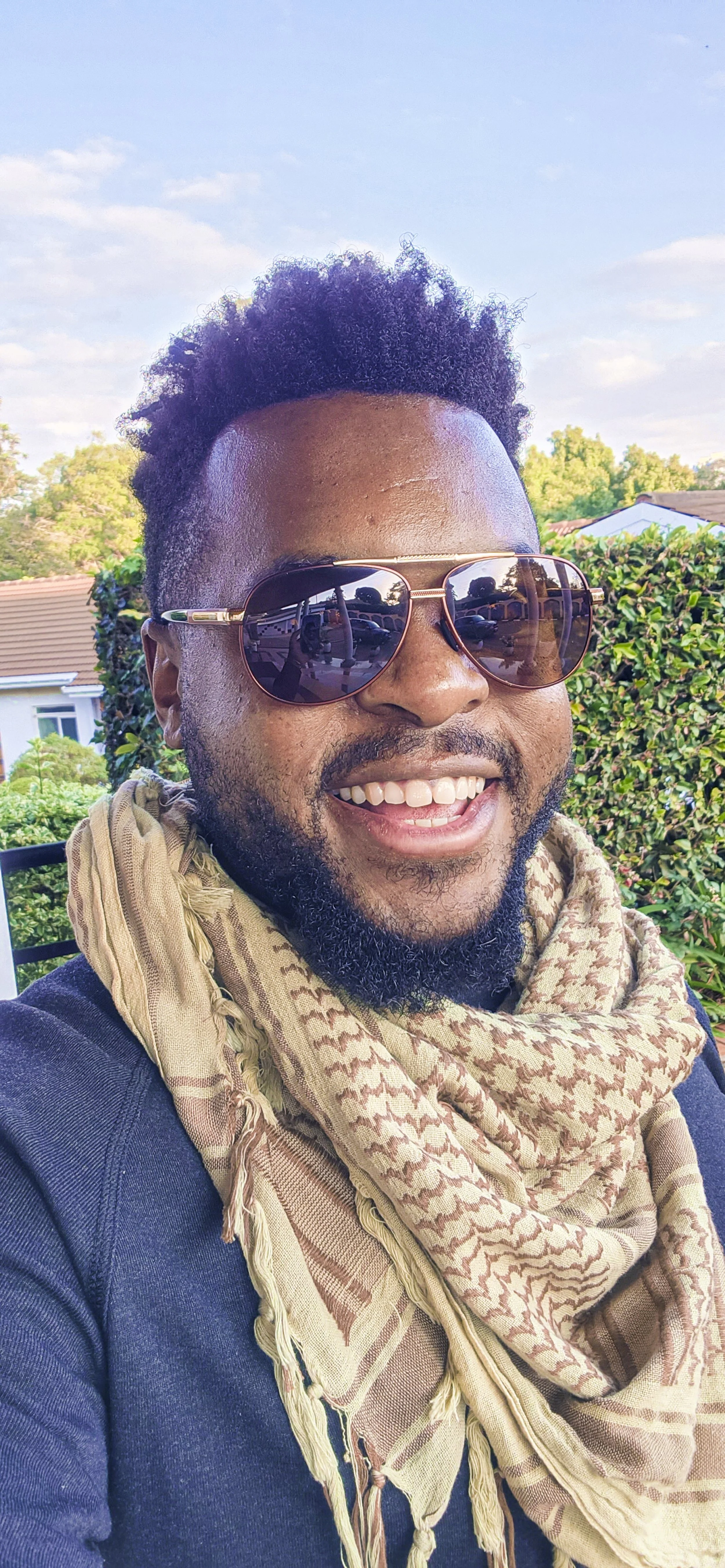 A smiling man wearing sunglasses and a patterned scarf outdoors with trees and houses in the background.