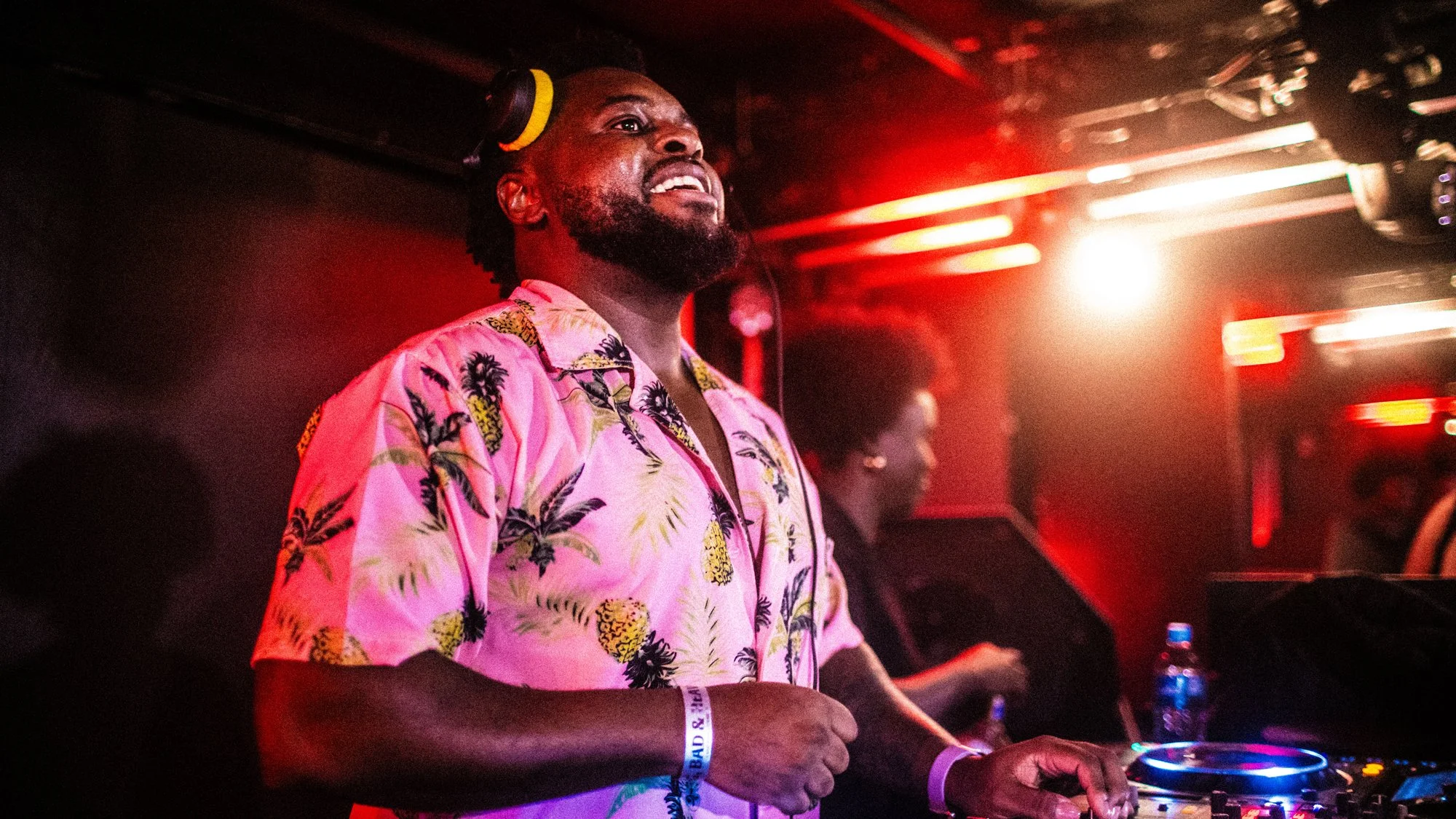 A DJ wearing a pink Hawaiian shirt with pineapple and palm tree designs, operating a DJ mixer in a club with red and orange lighting.