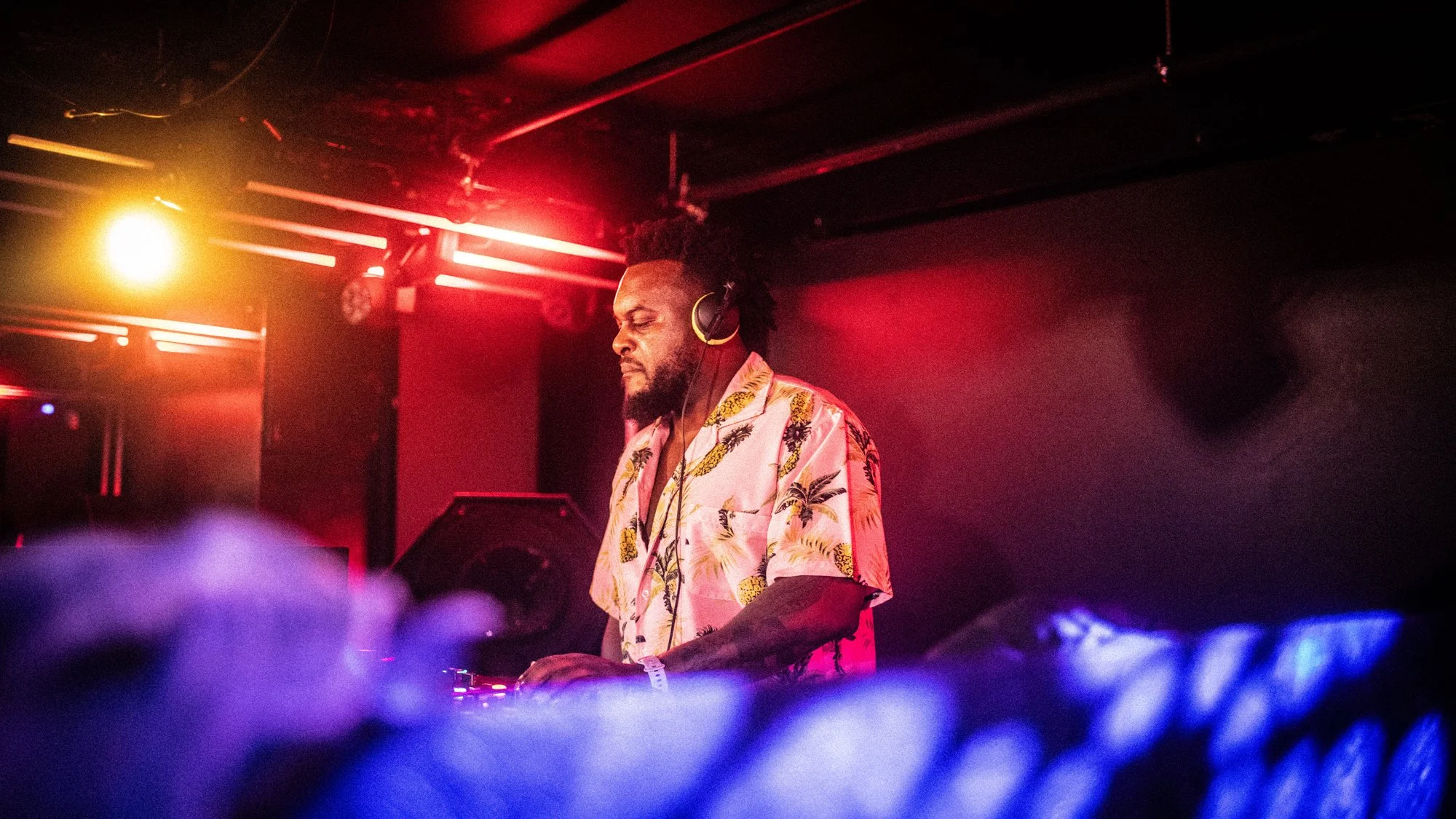 DJ performing at a club, wearing headphones and a colorful Hawaiian shirt, with colorful lighting and a dark background.