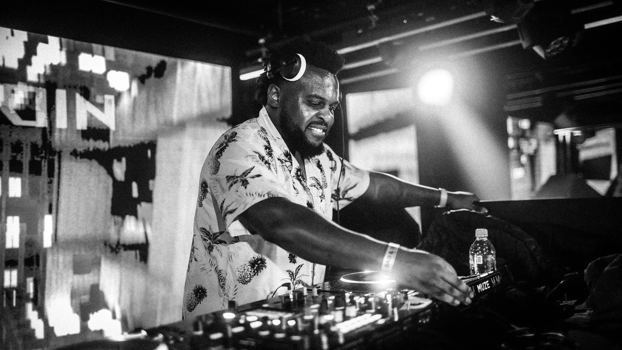 A DJ with headphones in a tropical shirt, smiling and operating DJ equipment in a dimly lit nightclub with a water bottle on the table.