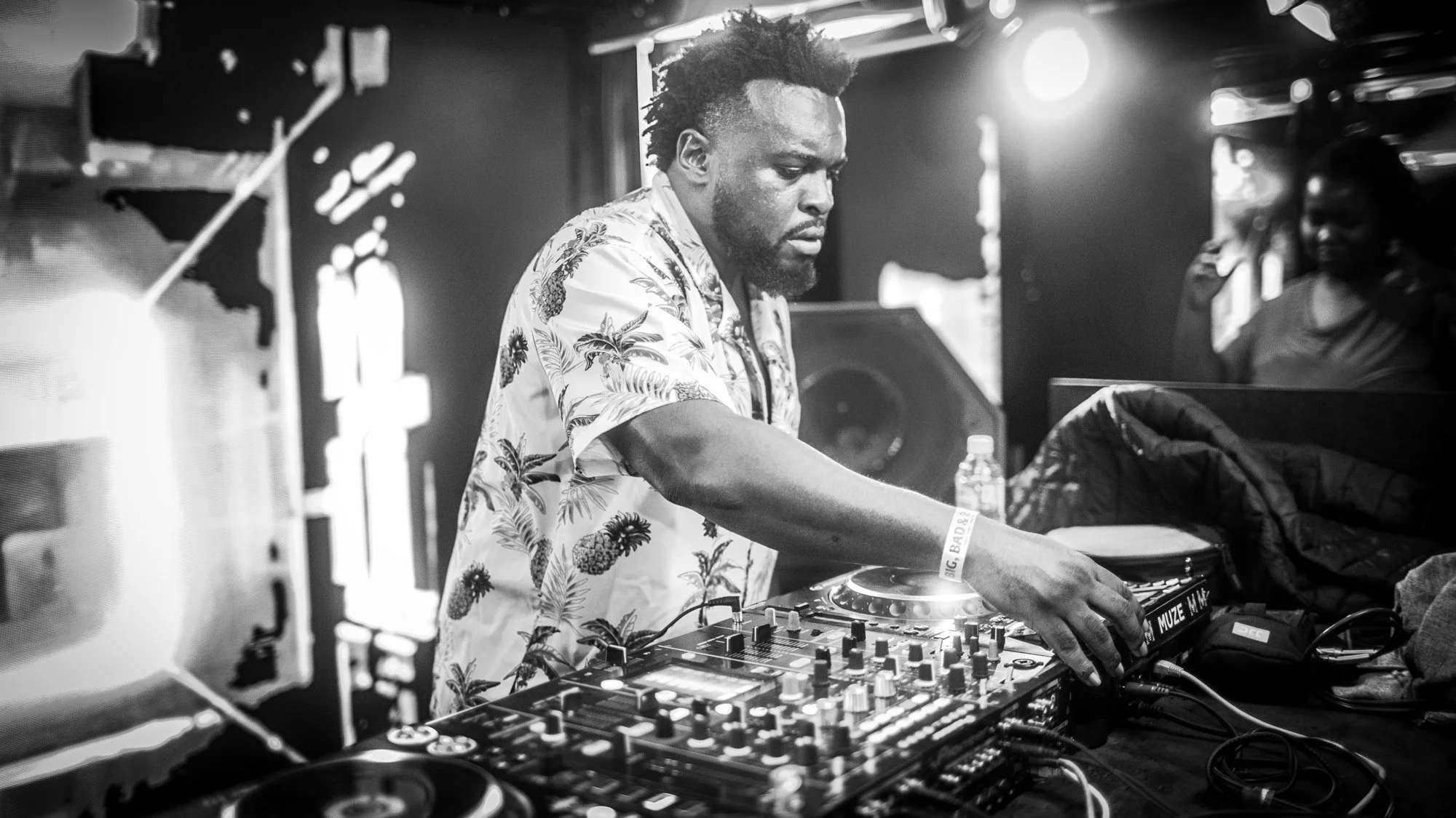 A DJ with curly hair wearing a floral shirt, mixing music on a DJ controller in a dimly lit venue, with a woman watching in the background.