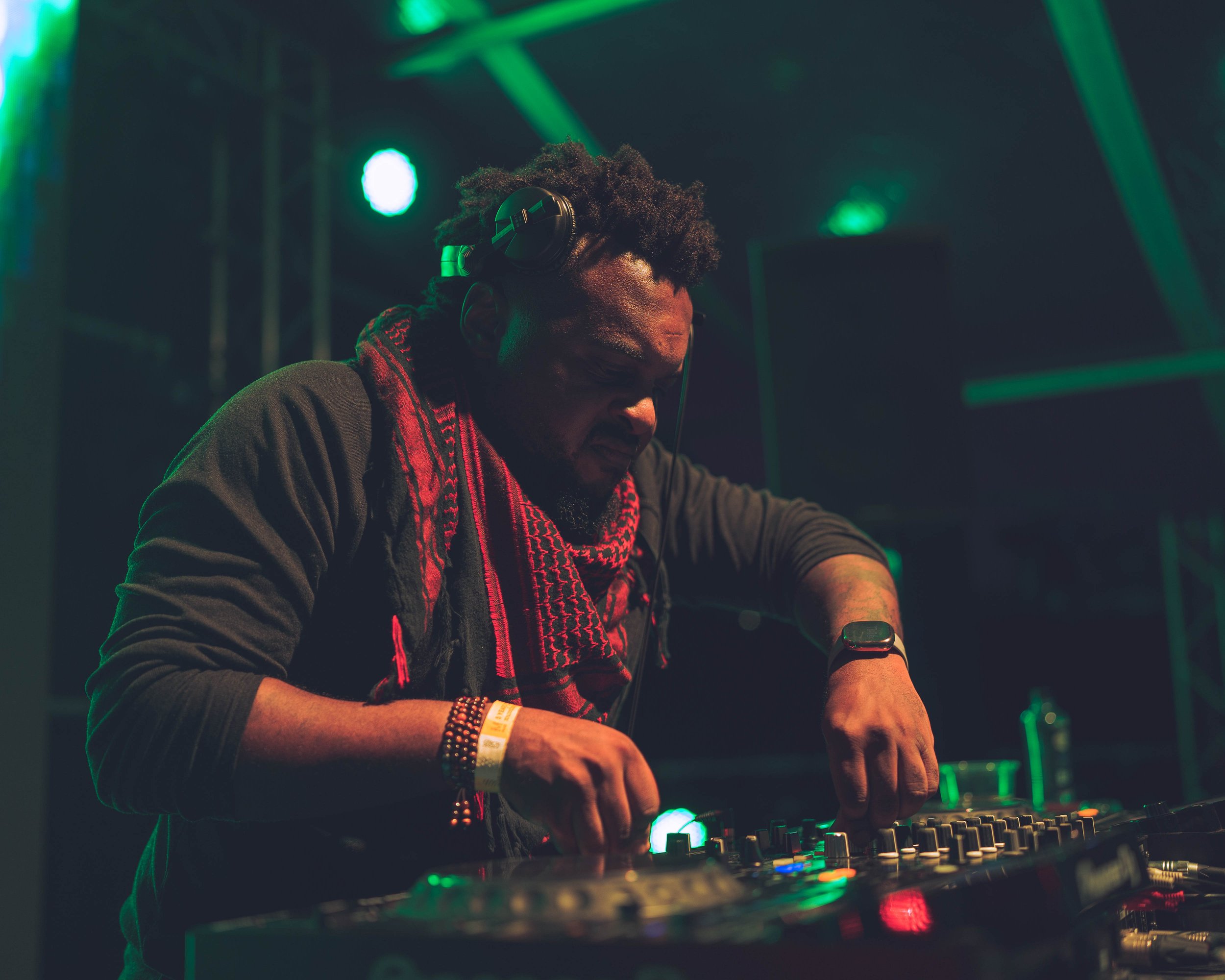 A DJ wearing headphones and a red and black scarf is mixing music on a professional DJ controller under green stage lighting.